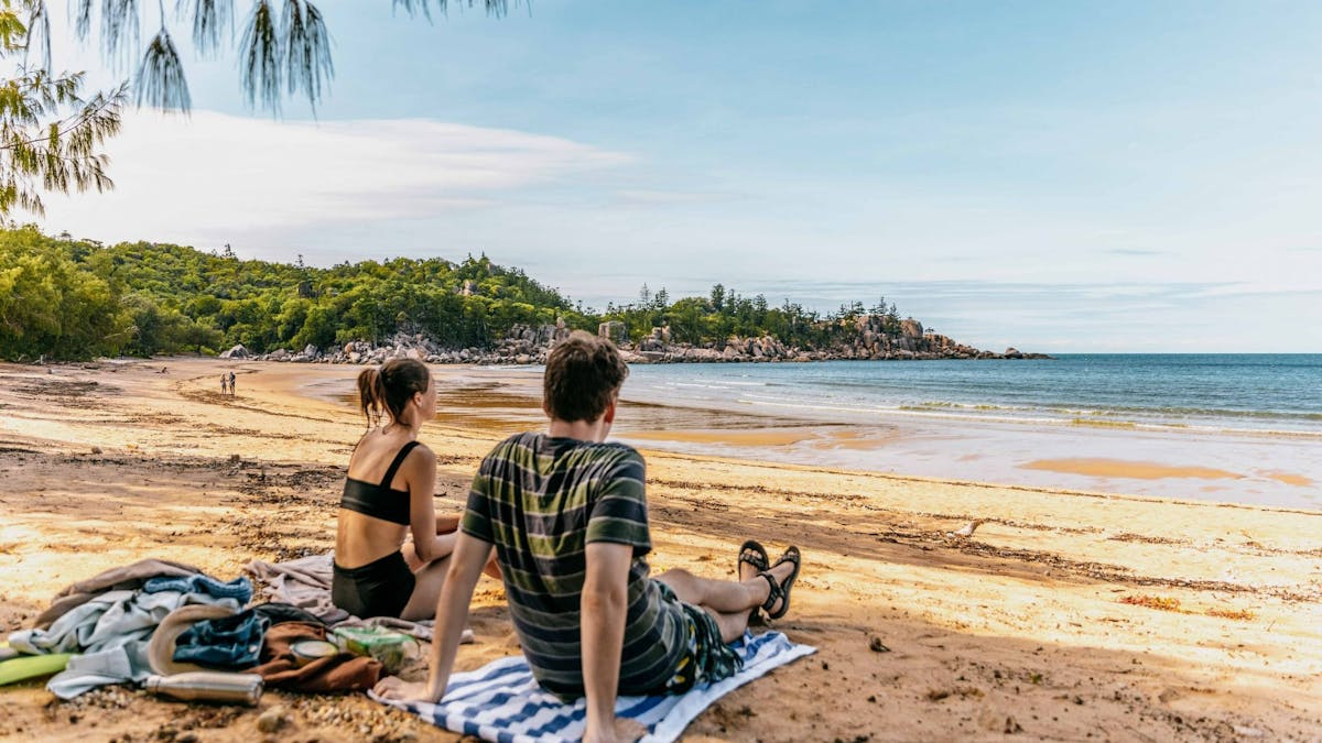 Two people sitting on towels on the beach looking out at a bay.
