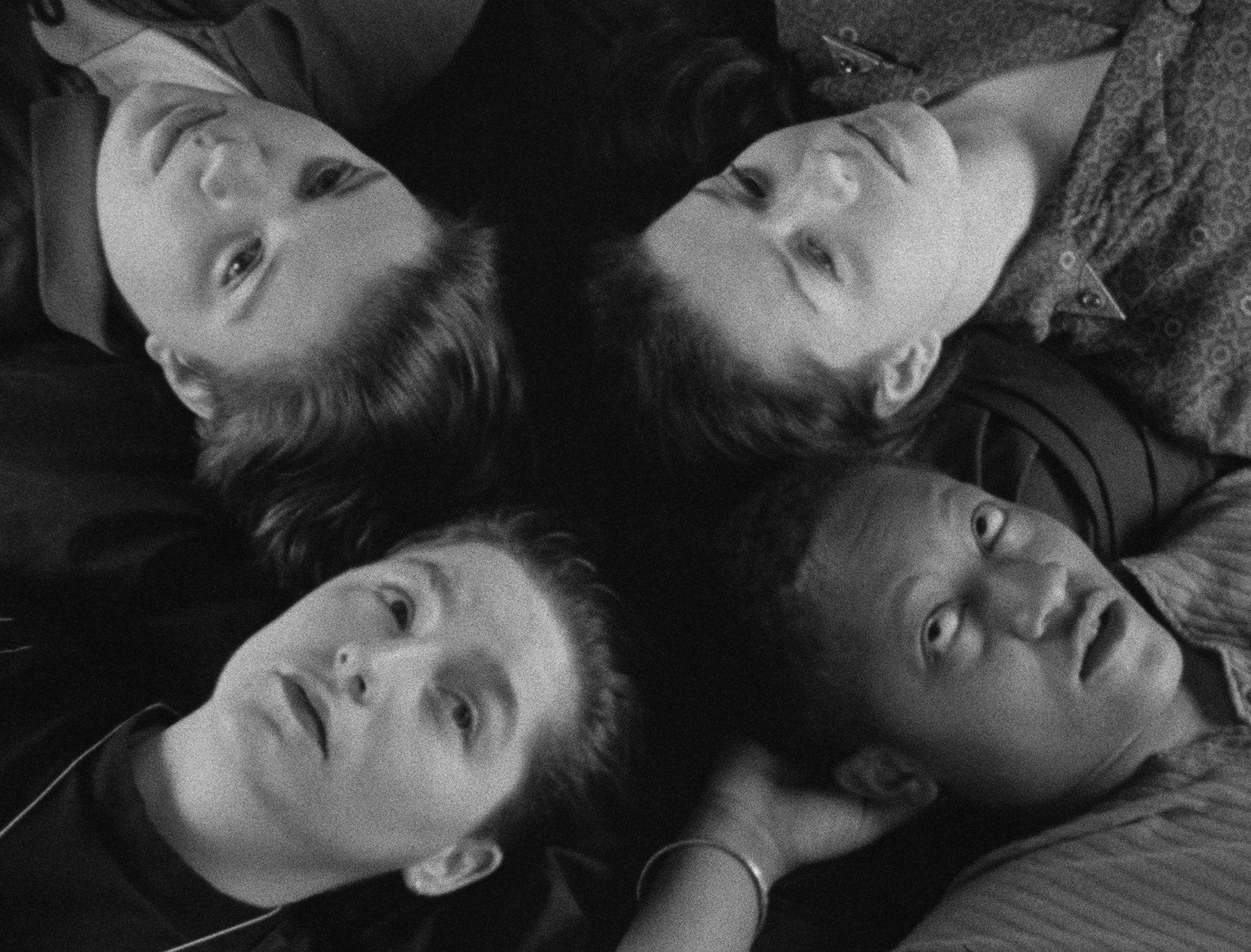 Four people lying in a circle, heads close together, looking contemplative in black-and-white.
