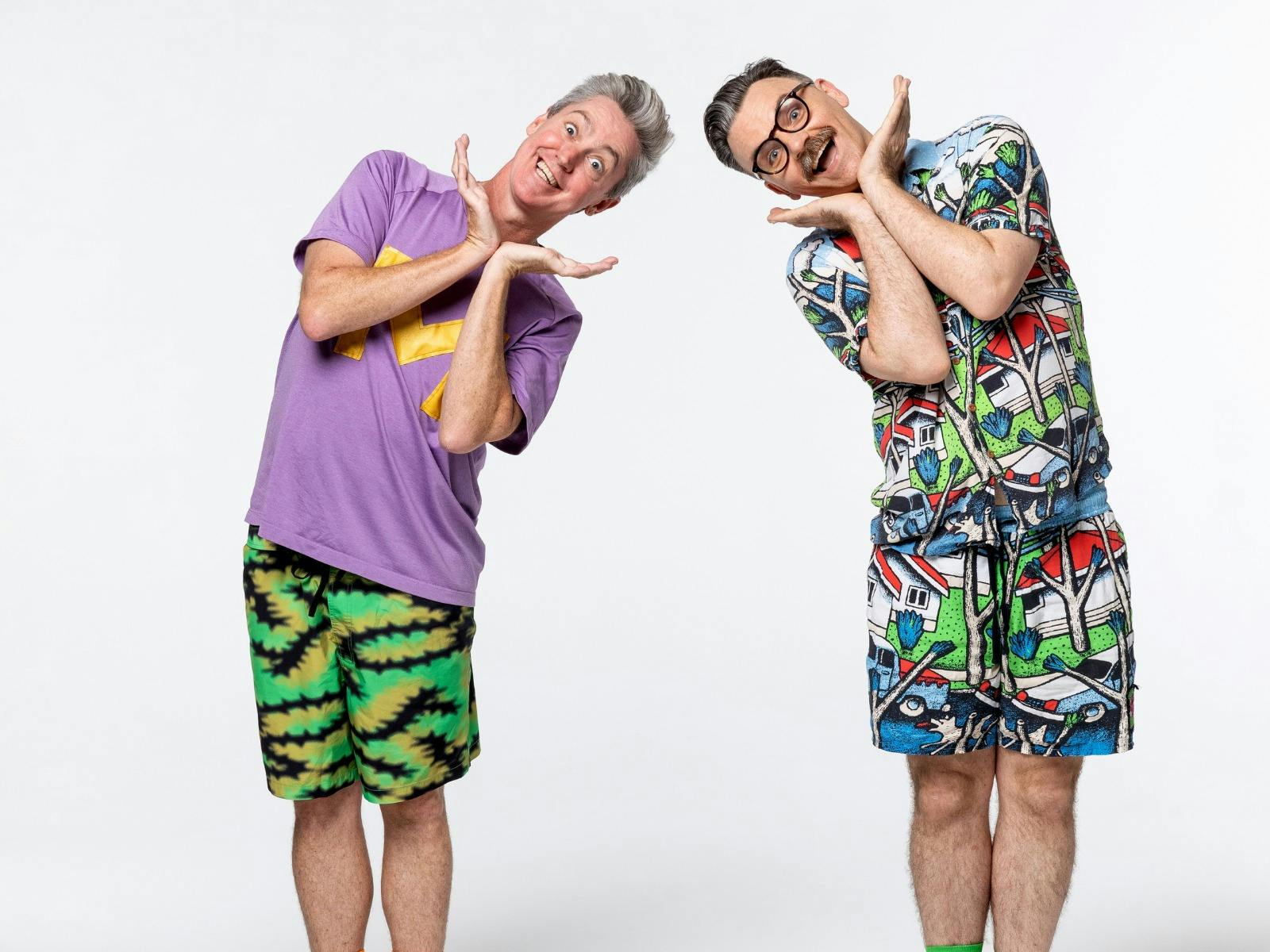 Two men in colourful clothing stand with eccentric facial expressions looking at the camera