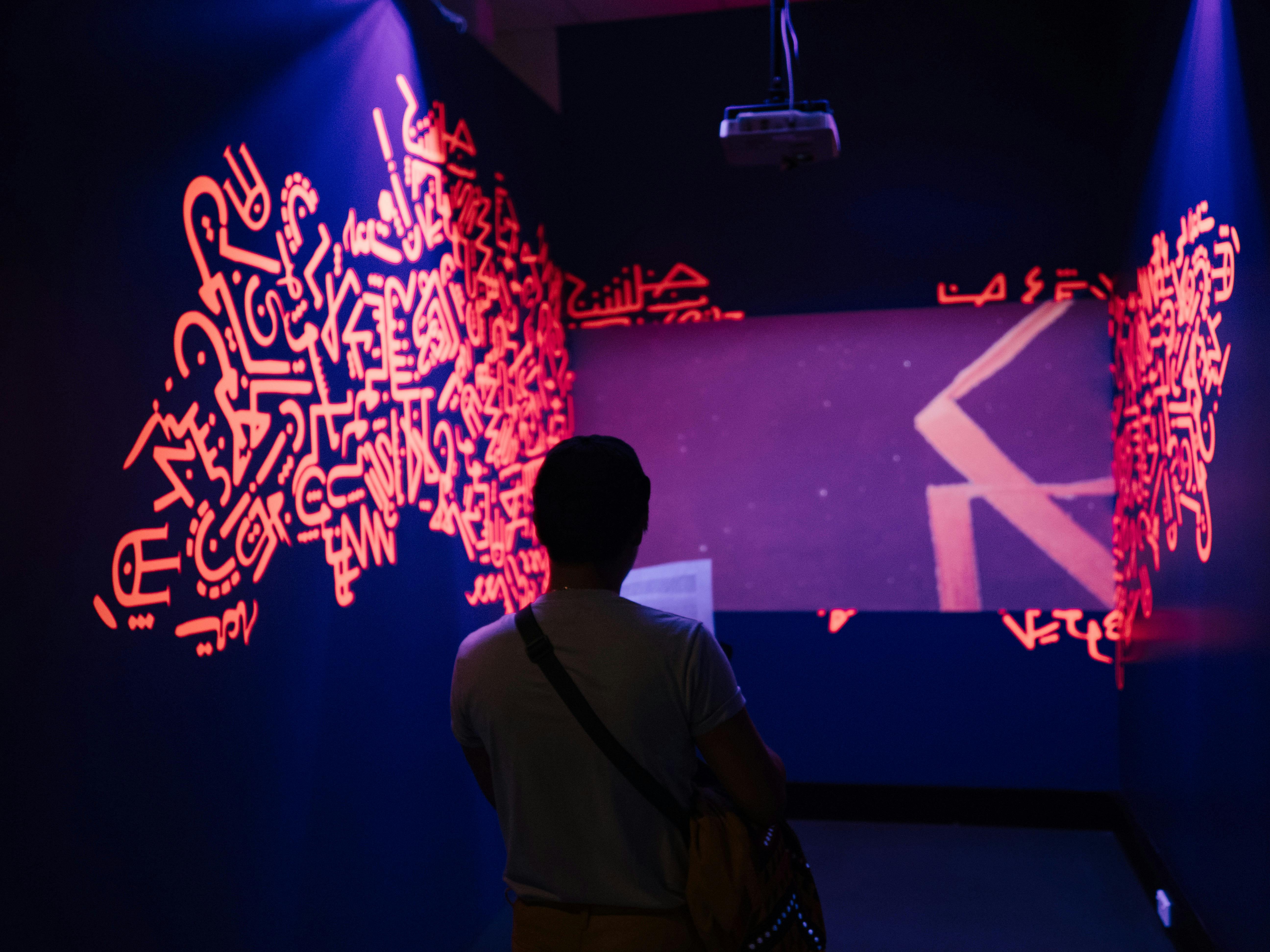 A photo of a gallery goer viewing Alia Ali's video work projected onto the wall with vinyl artwork