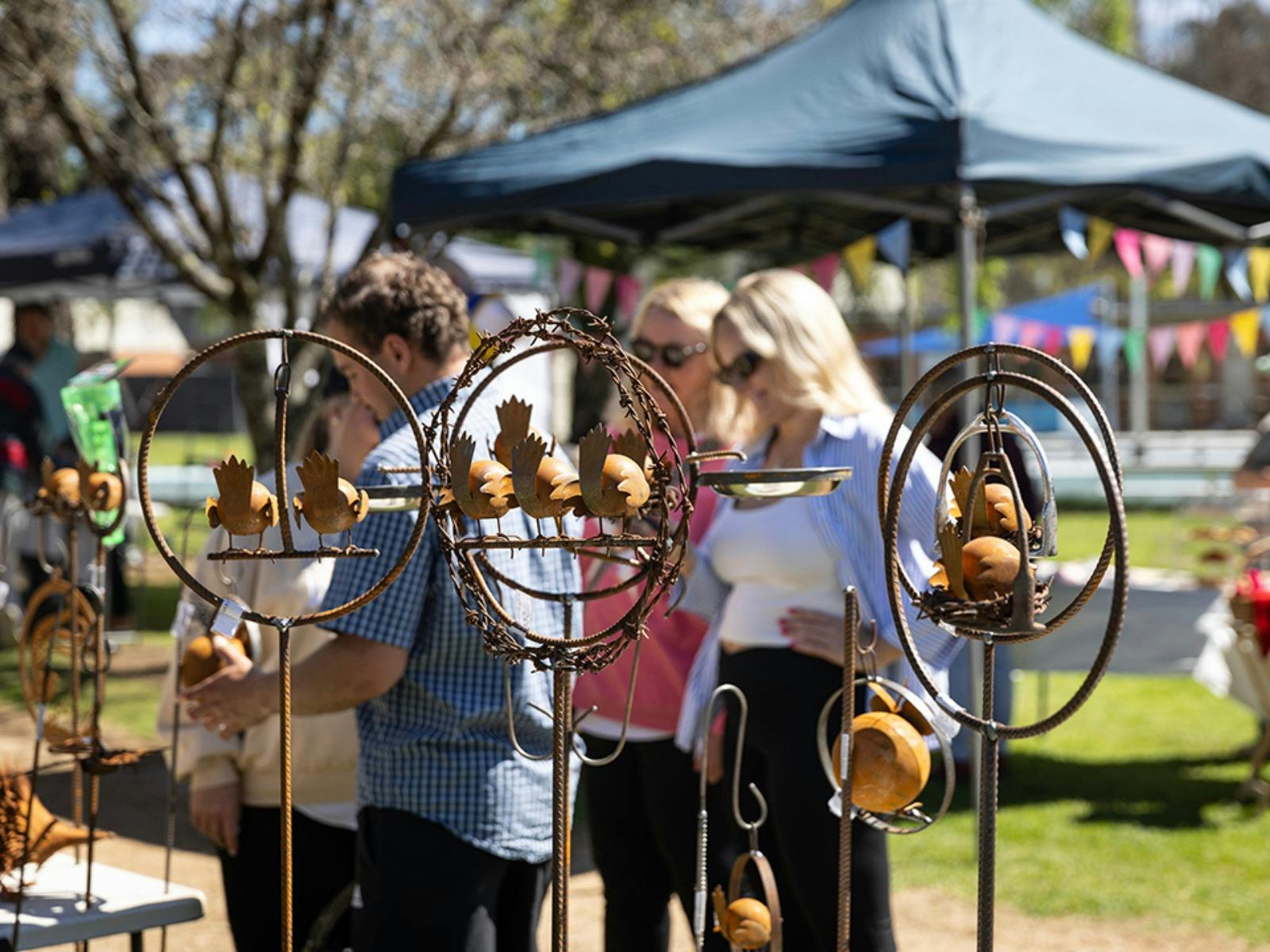 People browsing stalls under shady trees at a sunny outdoor market.
