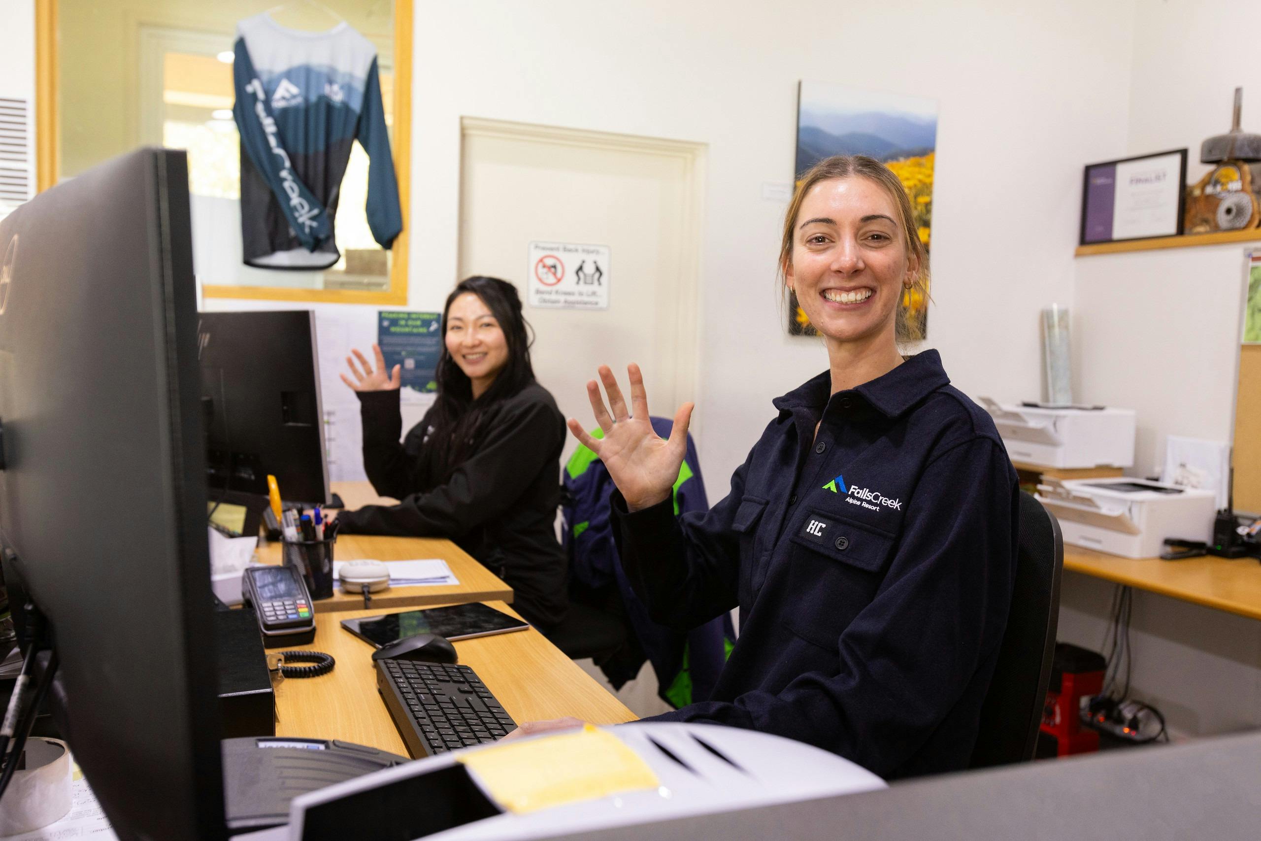 Two young female staff waving towards the camera as they sit at the information centre front desk.