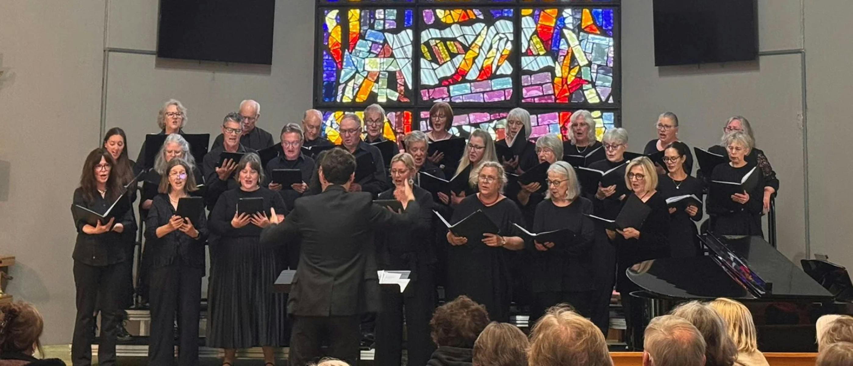 A group of singers dressed in black sing in front of a stained glass window.
