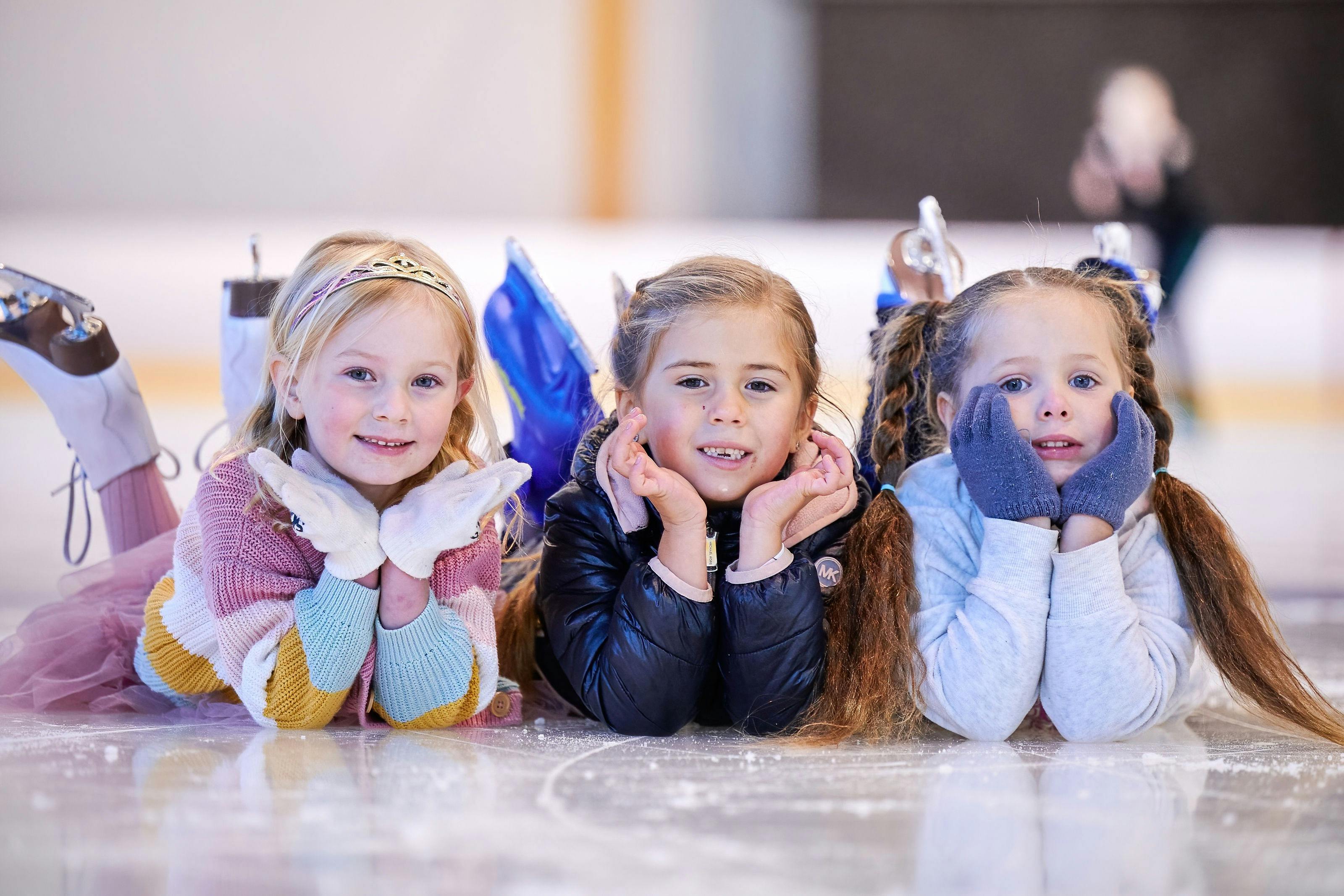 3 young preschool aged girls lying on the ice surface smiling at the camera.