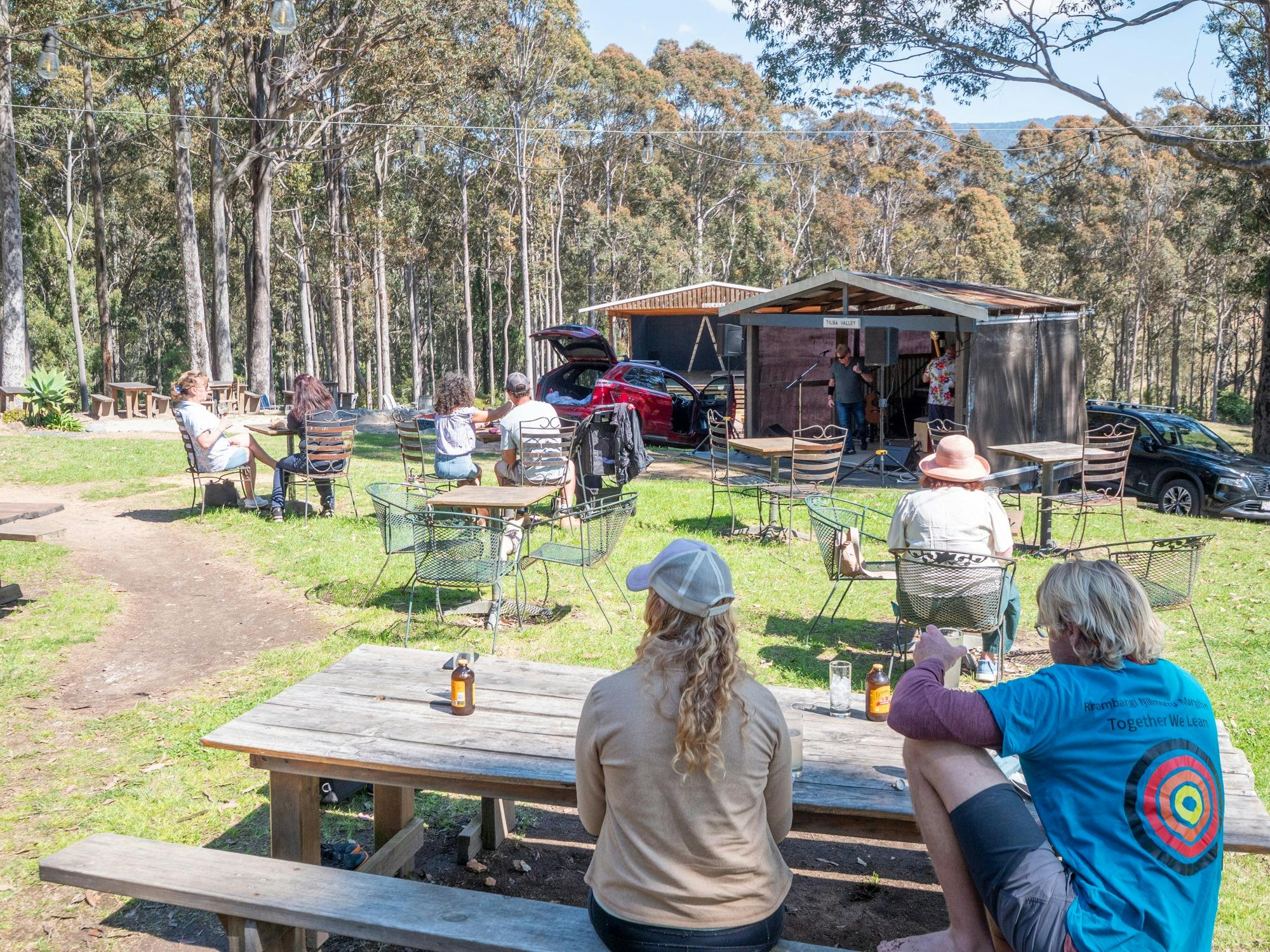 Two people sitting at a picnic table overlooking a music stage set against a lush, forest.