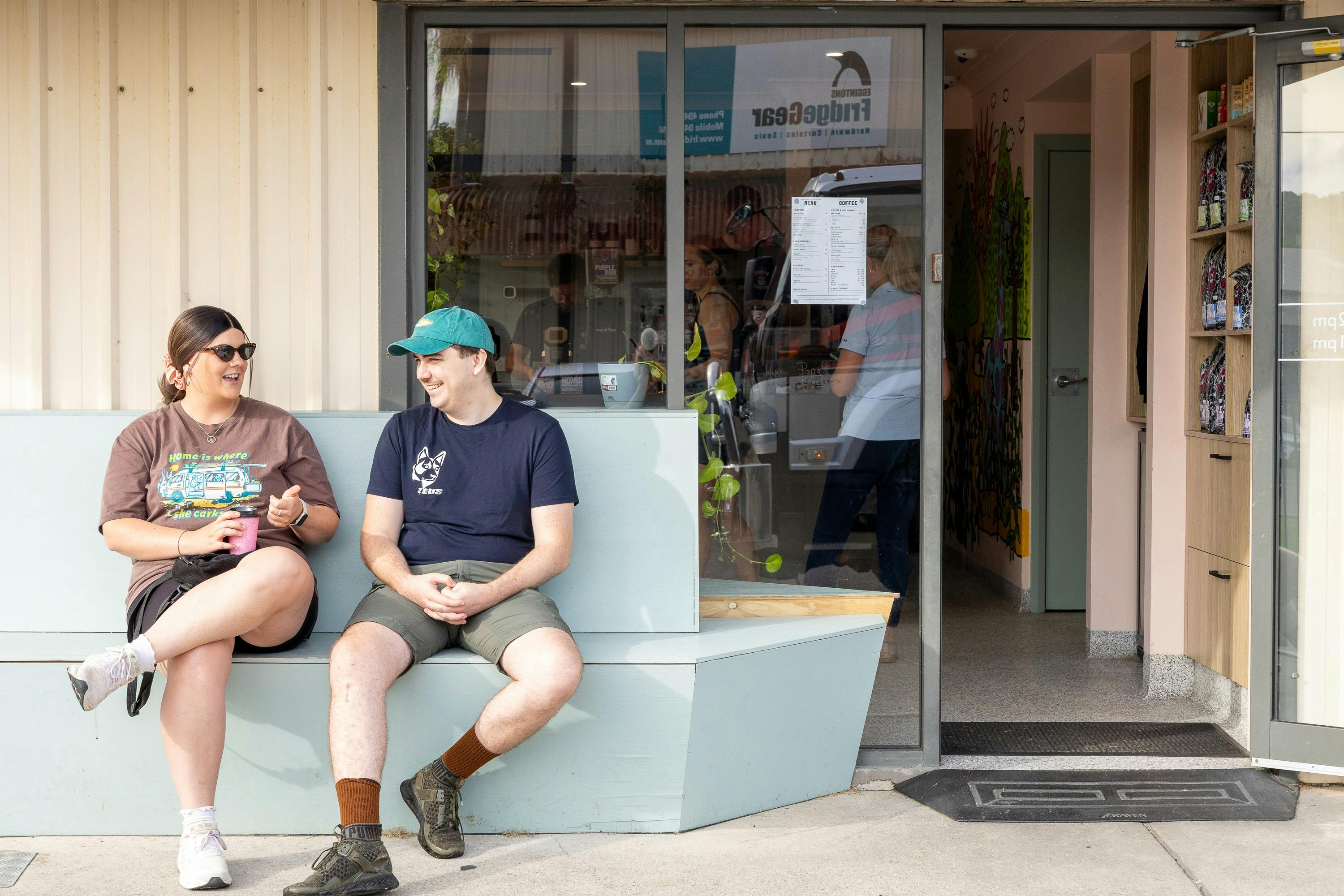 A couple sit at the bench outside the front door. You can see inside the café through the window.
