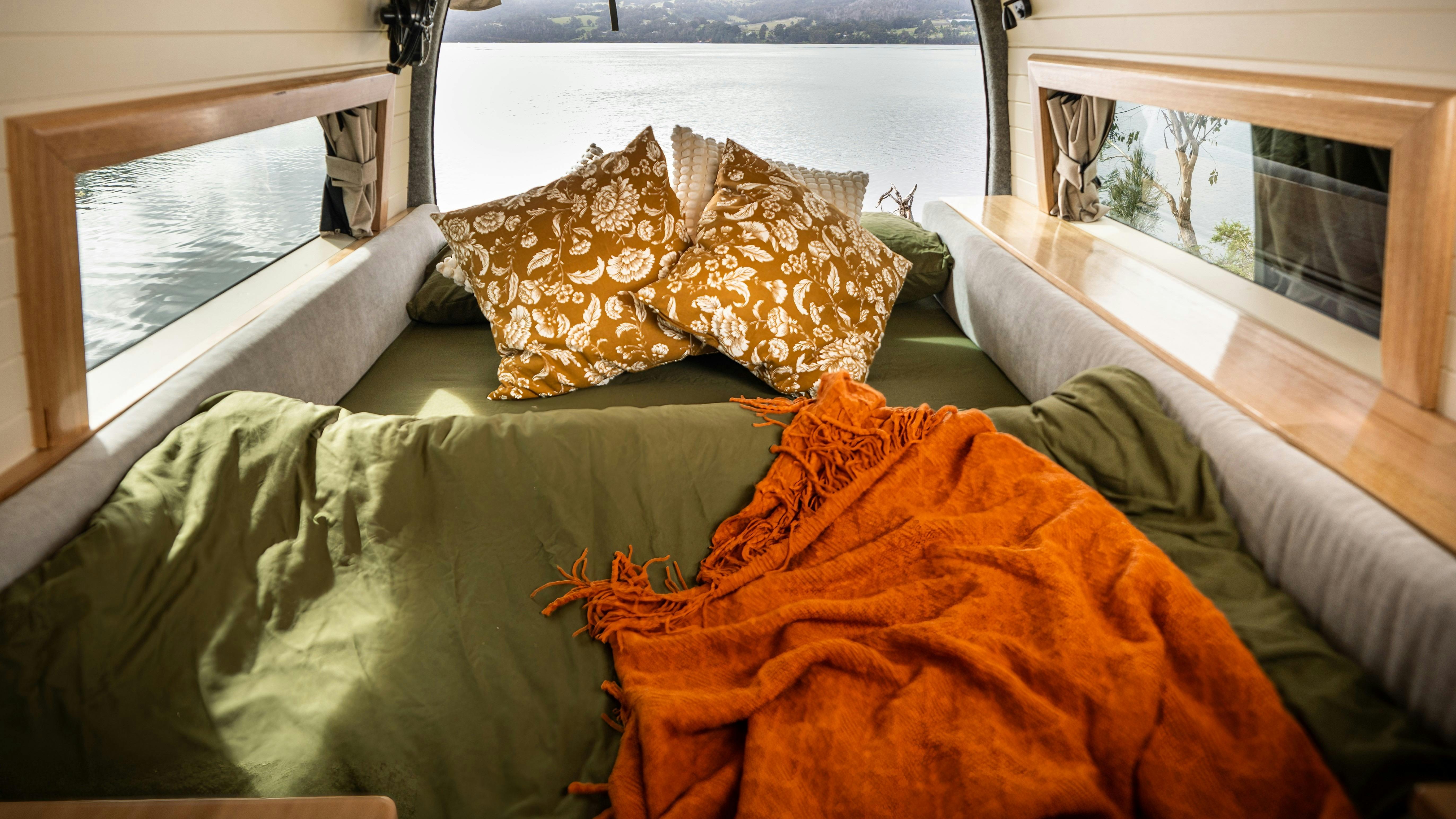 Campervan sleeping area with generous bed size and scenic Tasmanian lake in background.