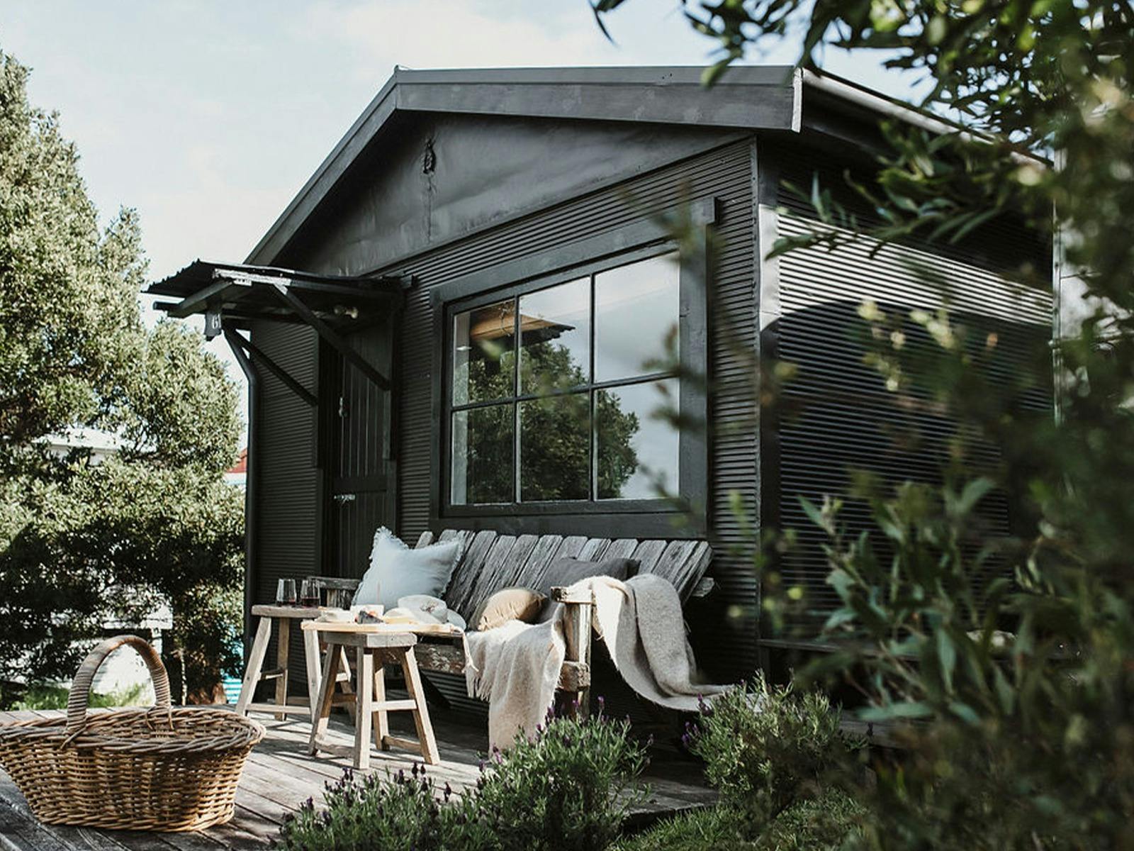 A black shack with a large picture frame window. A bench and chairs sit on the deck in front.