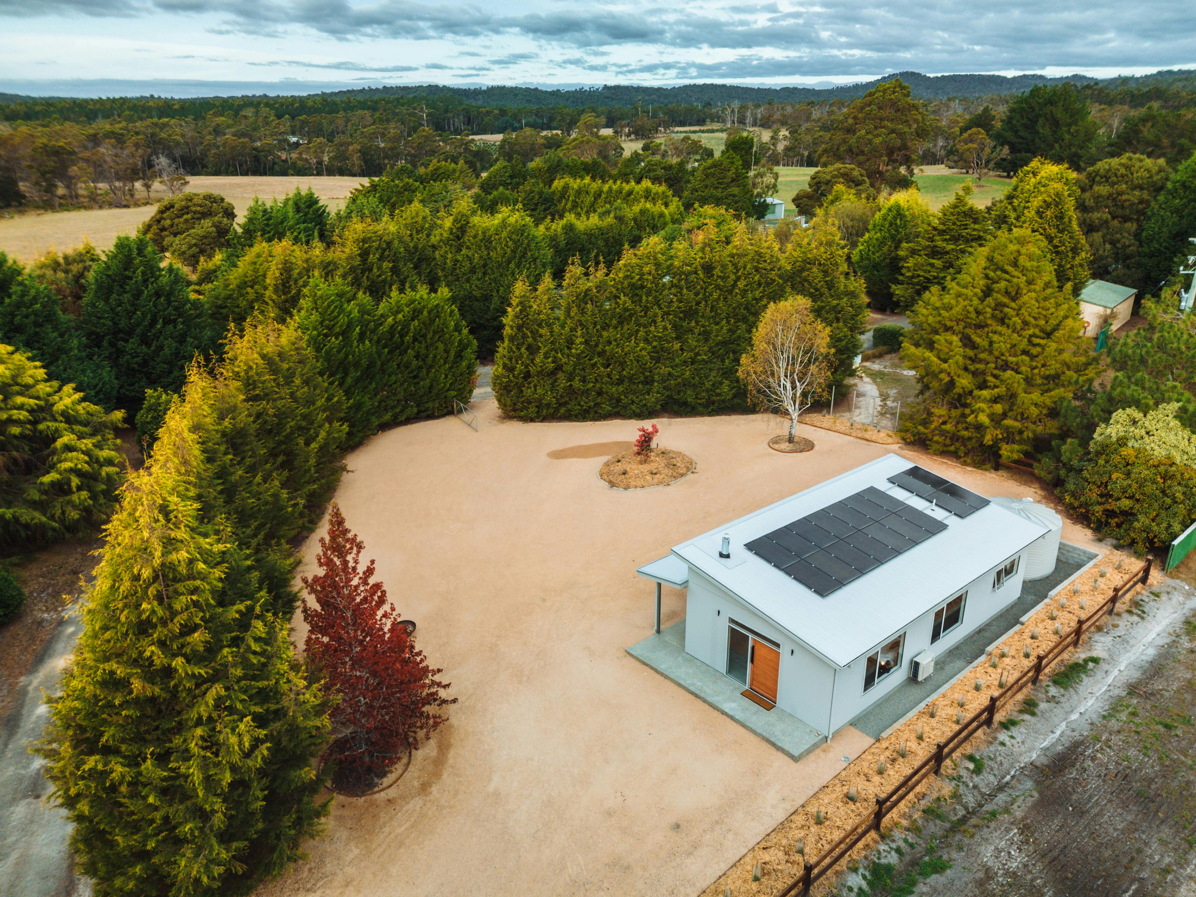White building with solar panels set within a parking area surrounded by pine trees and garden