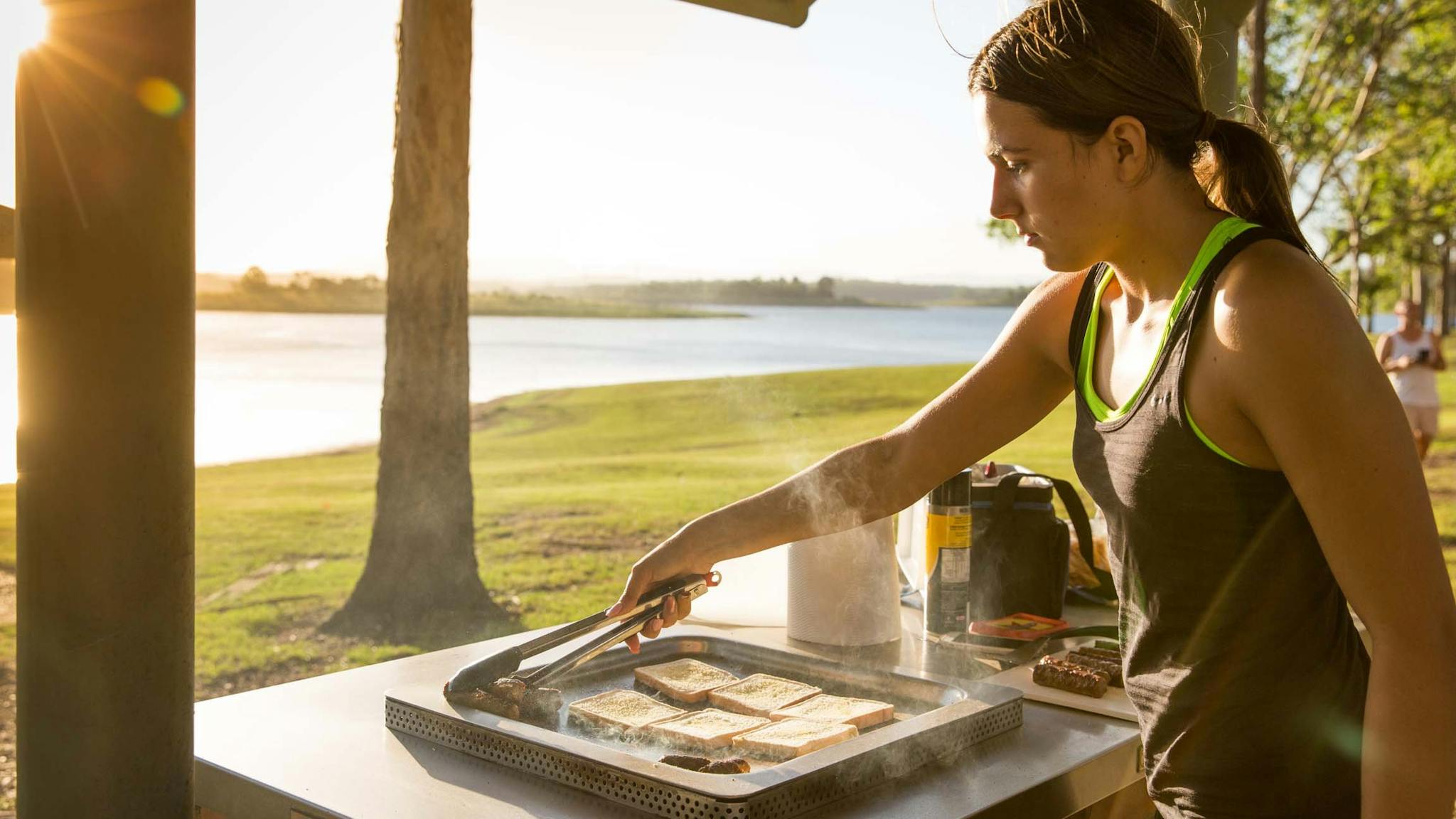 Woman flipping sausages with tongs on a barbecue under picnic shelter looking over grass and lake
