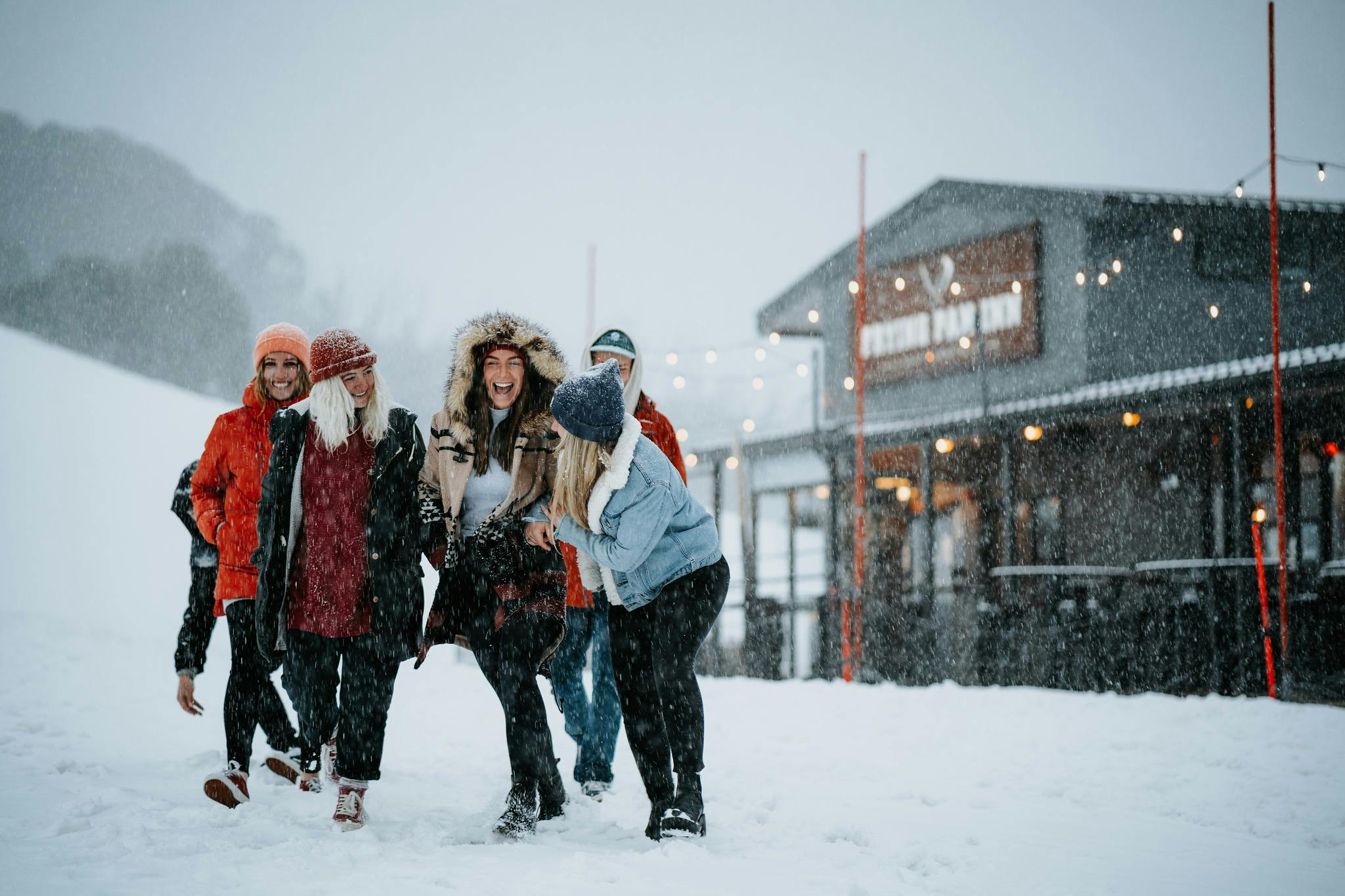 A group of six smiling and laughing friends walk in the snow away from a Falls Creek alpine venue