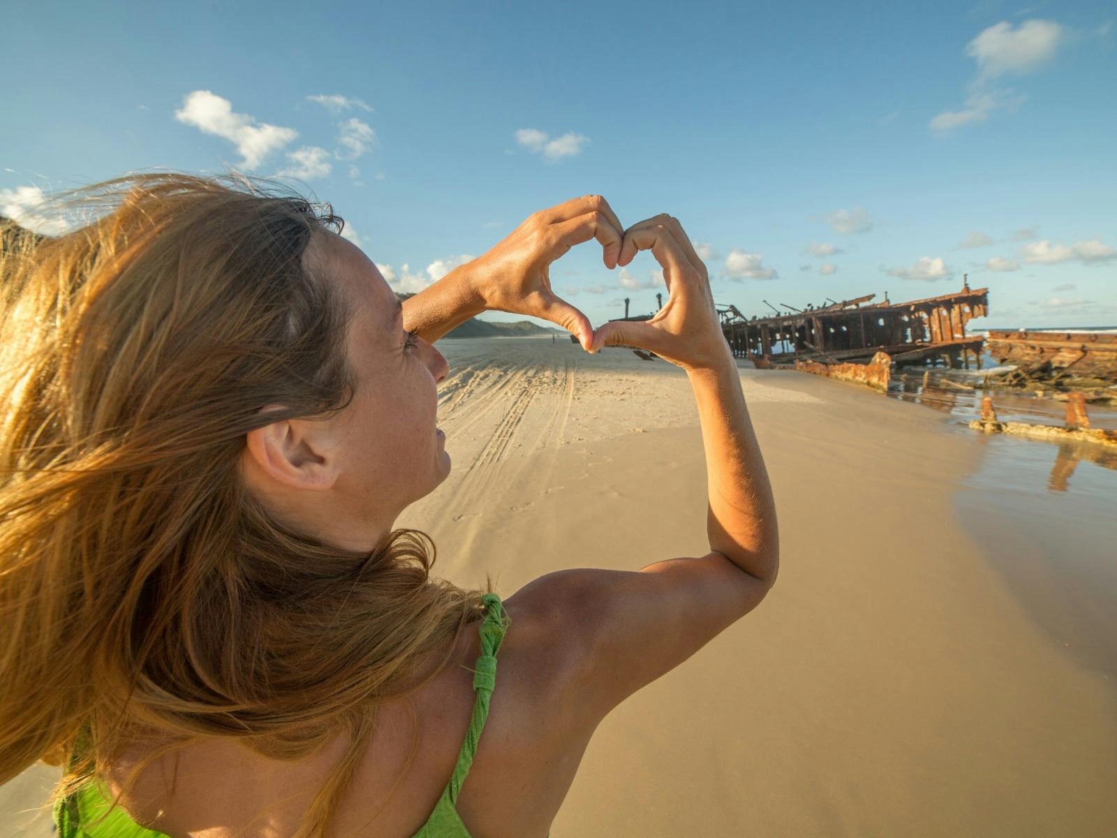 A young blonde woman holds her fingers in a love heart sign at a shipwreck on a sandy beach A young blonde woman holds her fingers in a love heart sign at a shipwreck on a sandy beach