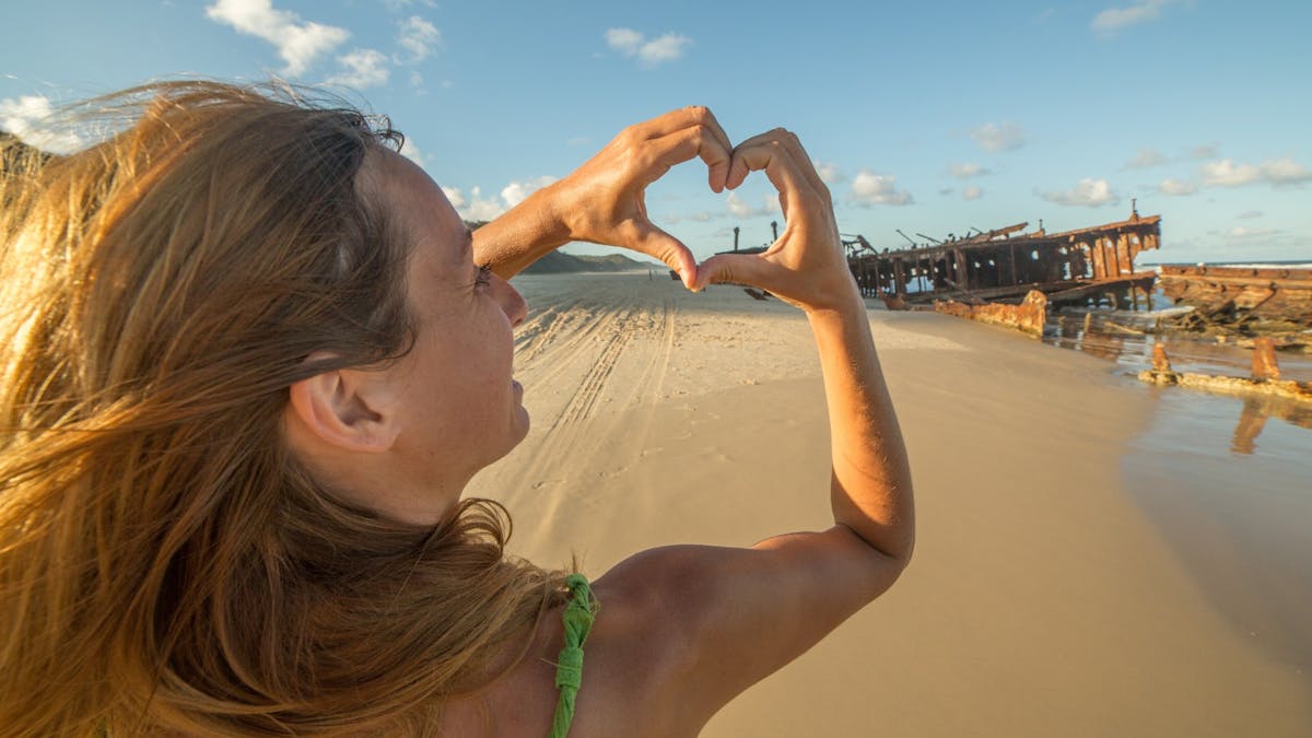 A young blonde woman holds her fingers in a love heart sign at a shipwreck on a sandy beach