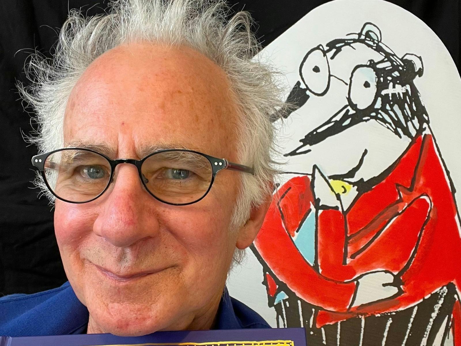 Man with glasses smiles beside a cartoon bear in a red jacket.