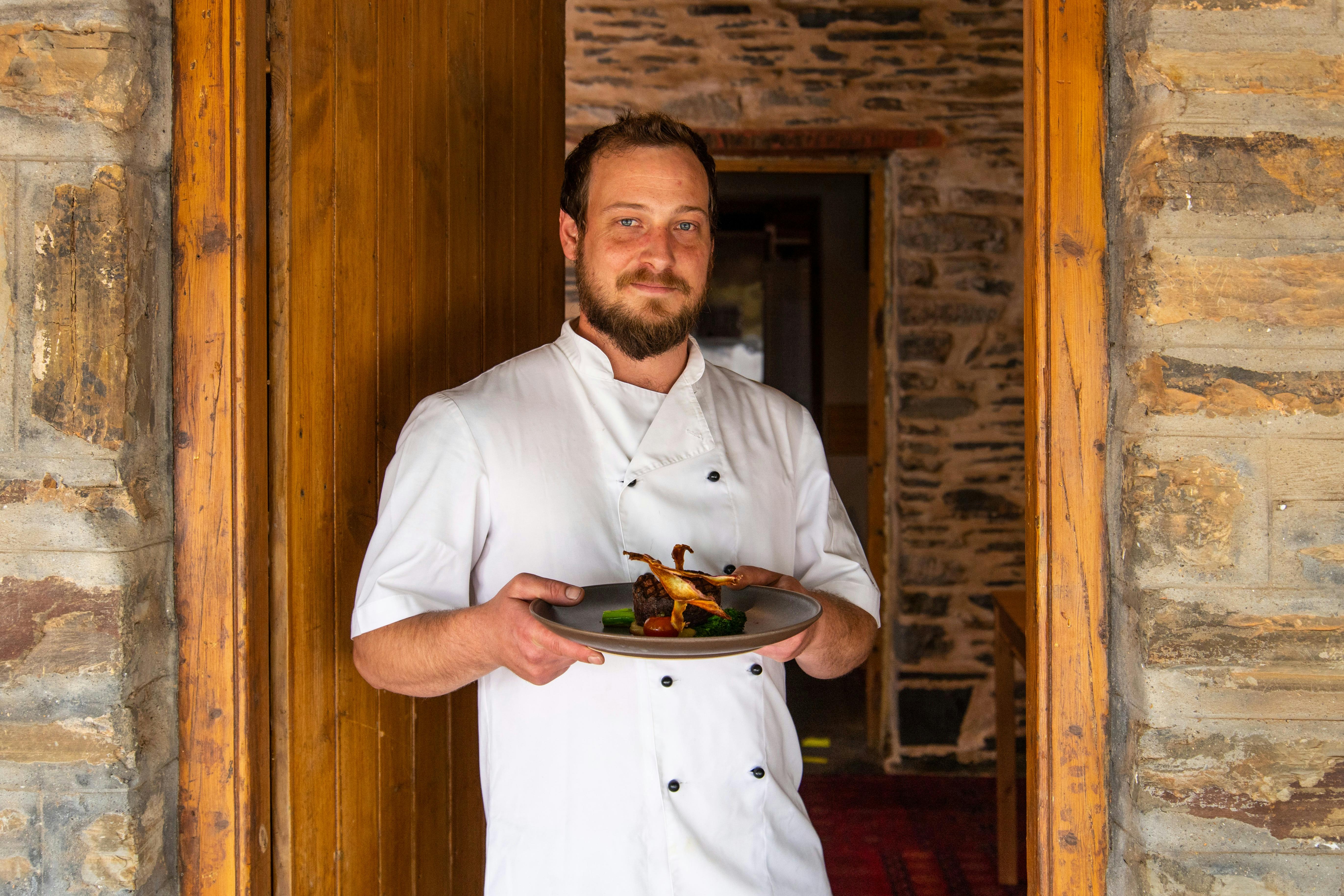 Reillys' Head Chef Luke Smith dressed in white chef uniform holding plate in front of heritage door