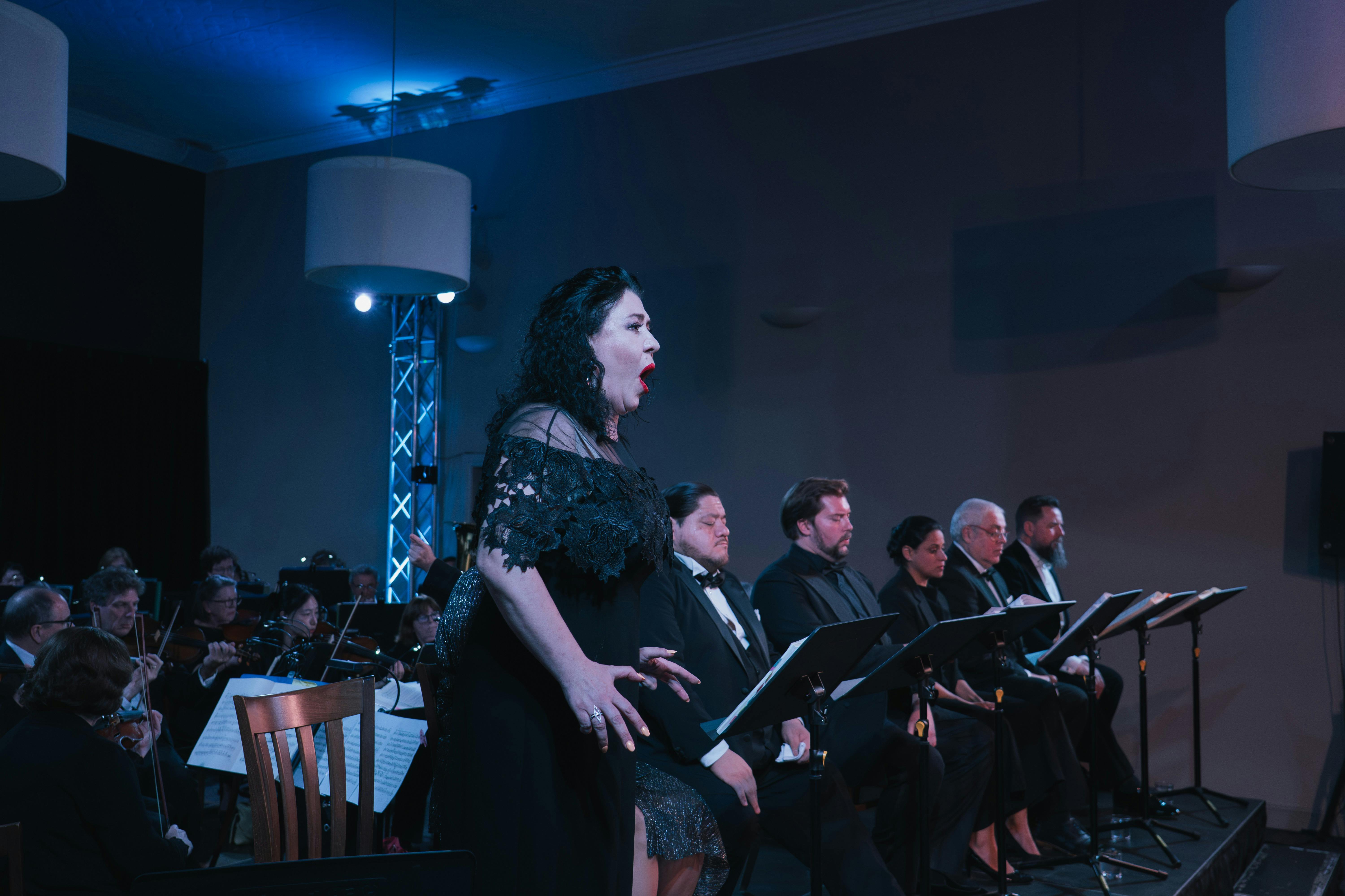 A singer in a black dress singing with other singers seated behind her
