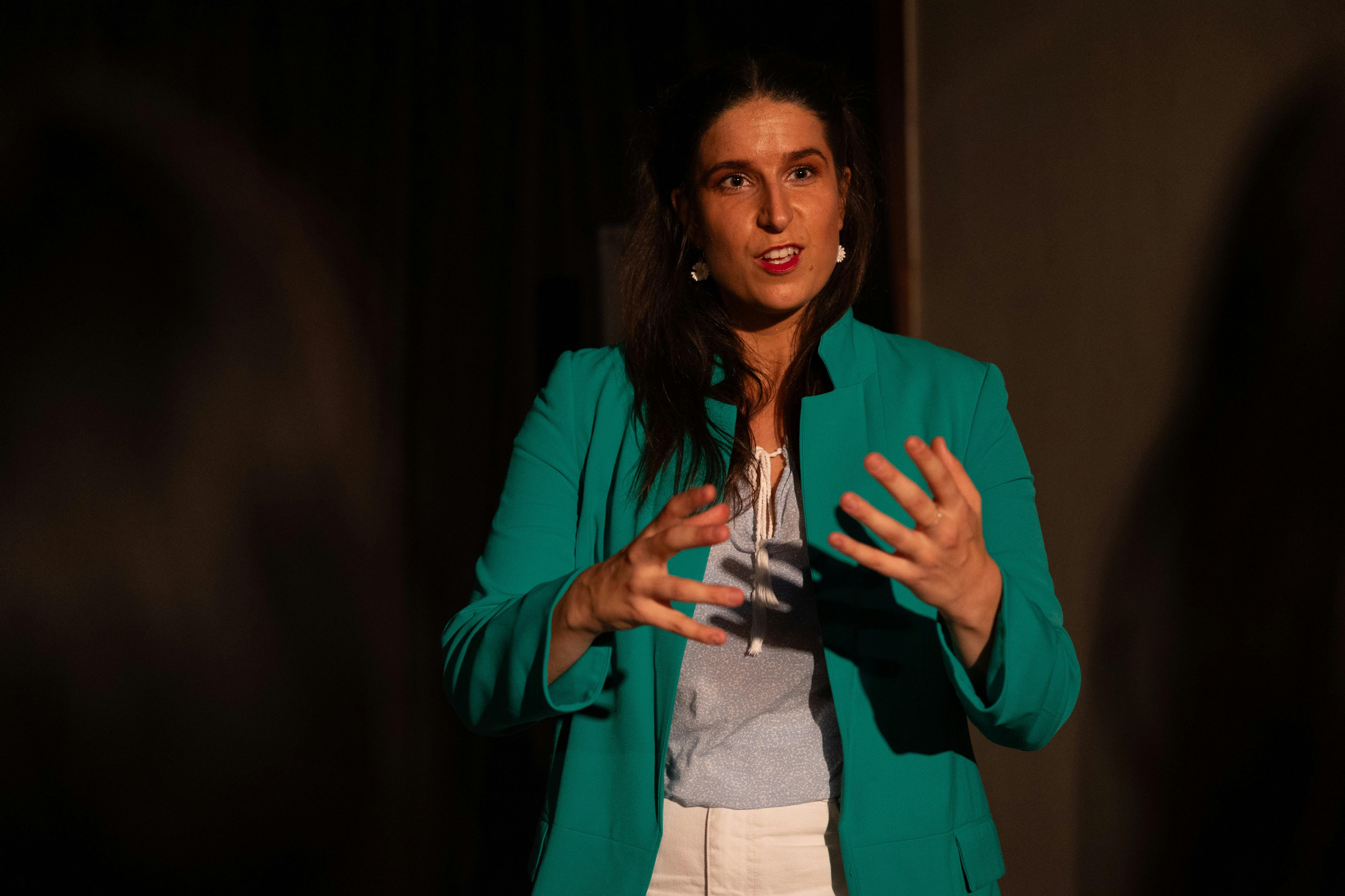 A woman storytelling in a green blazer, blue shirt and raised arms to express the story.