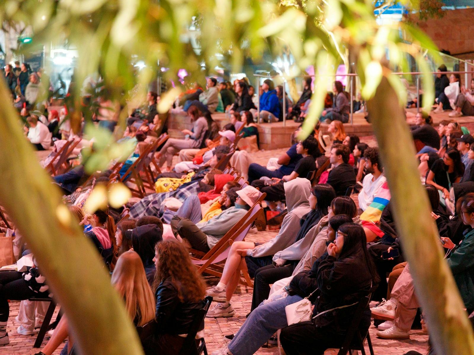 a crowd of people sit on deck chairs in the evening, watching something on the big screen