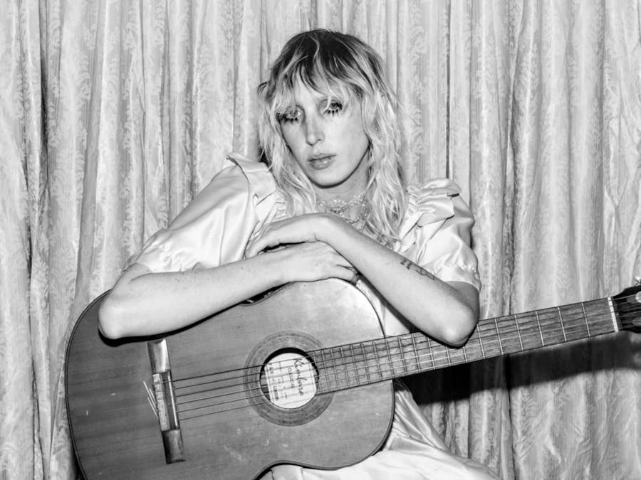 Babitha sits in front of a stage curtain holding her guitar
