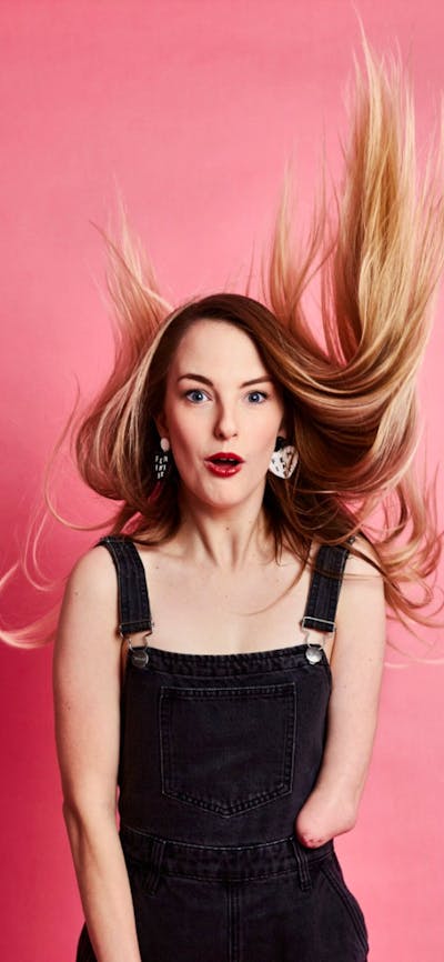 A woman in overalls standing in front of a pink background with her hair in motion
