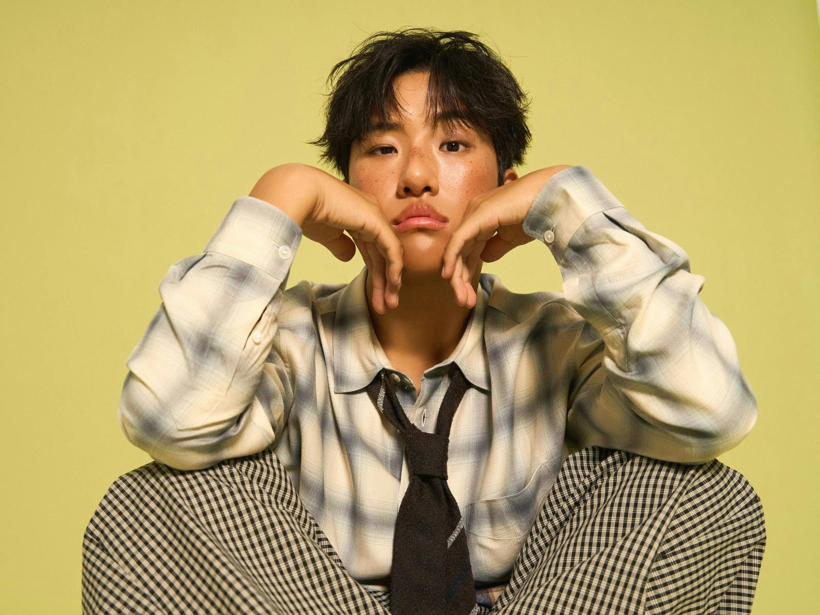 KINKY sitting down with his hands placed under his chin, with a yellow background