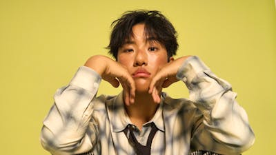 KINKY sitting down with his hands placed under his chin, with a yellow background