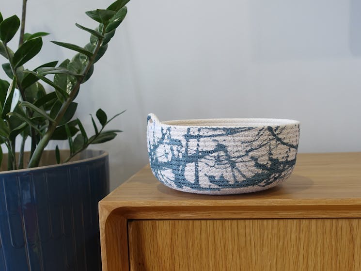 A blue and white handmade rope bowl on a sideboard, with a plant to the left