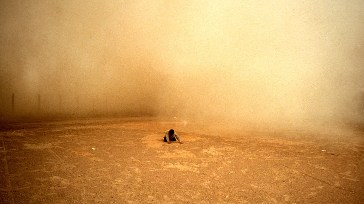 A person in the distance kneeling and looking downwards, surrounded by orage dust A person in the distance kneeling and looking downwards, surrounded by orage dust