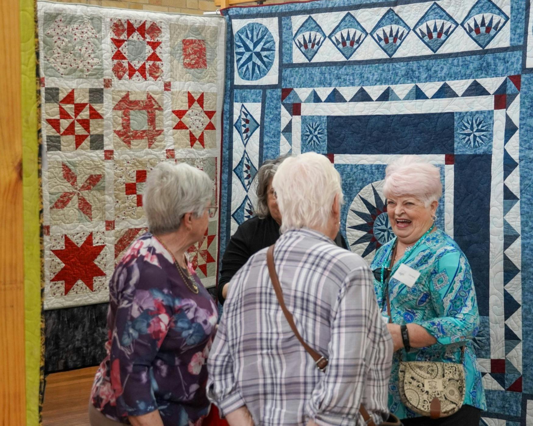 Ladies gathering together. One lady laughing. Standing in front of quilt display