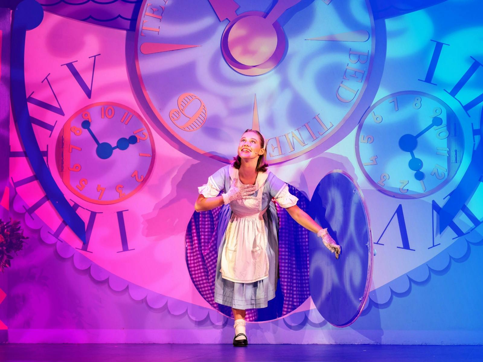 Performer dressed as Alice steps through a clock portal on stage, with colorful clocks behind her.