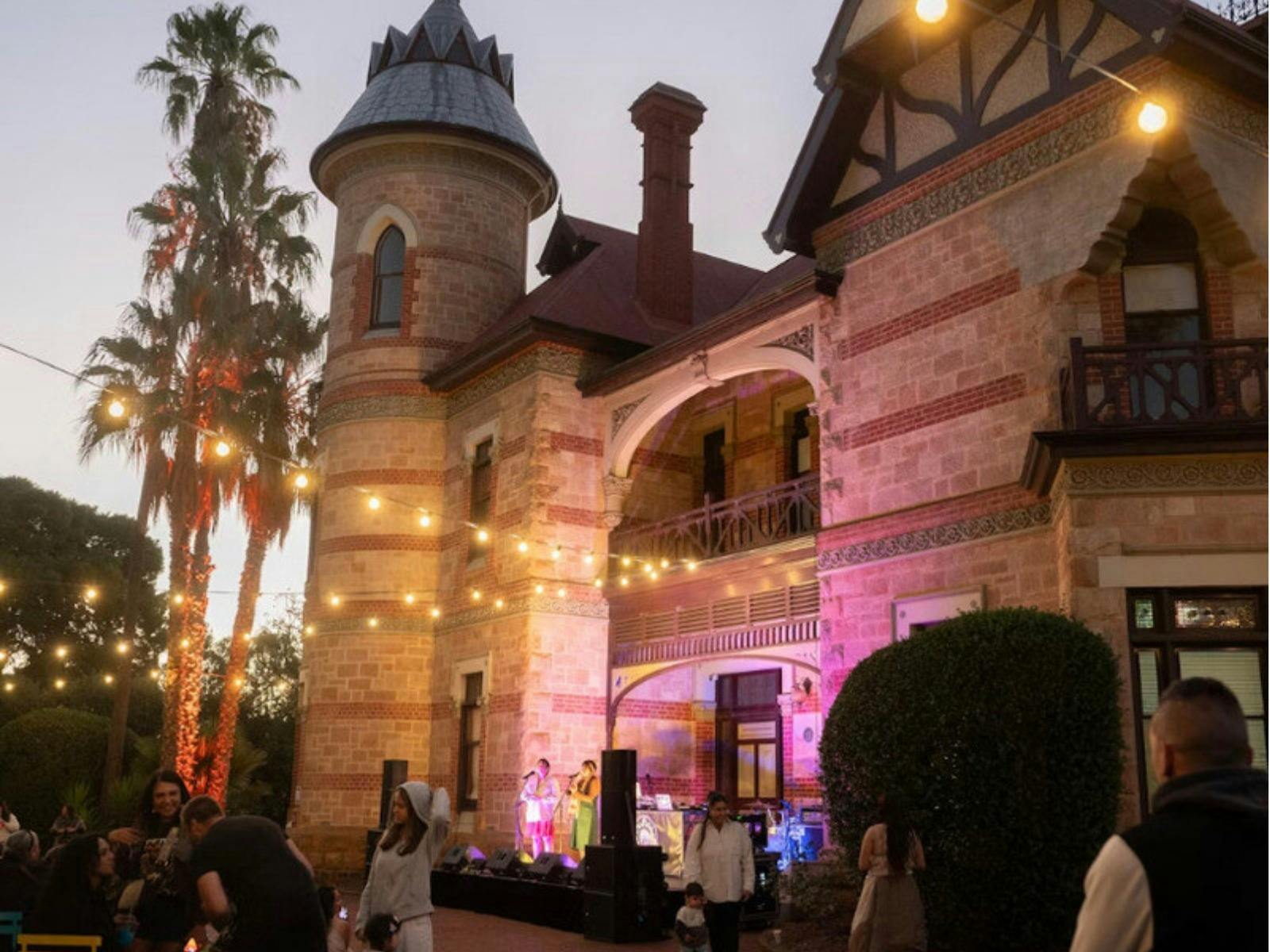 Historic mansion illuminated by string lights with a lively outdoor gathering at dusk.