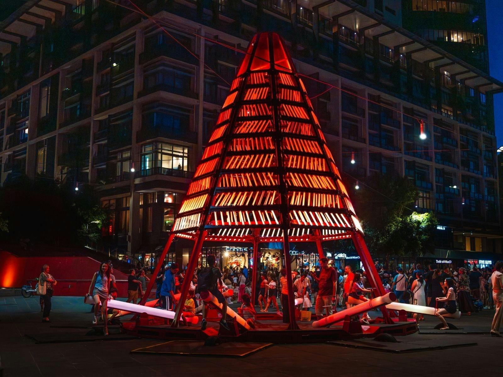 A large red sculpture stands prominently in the center of a bustling city