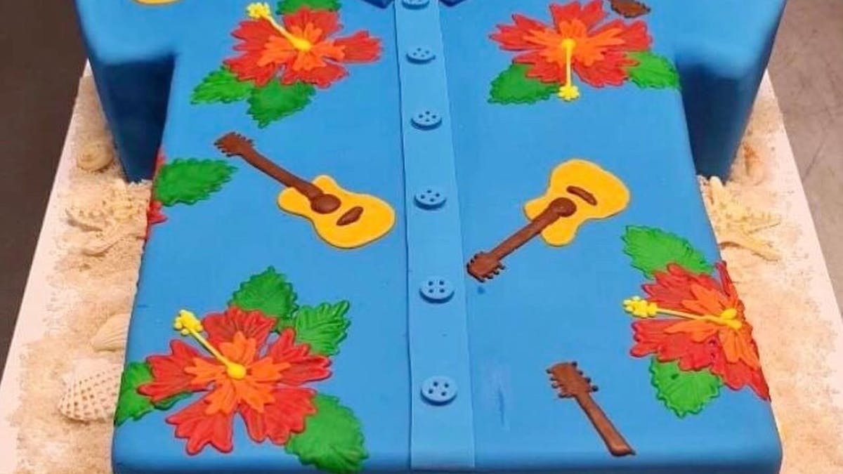 A cake made in the shape of a blue collared shirt with guitars and hibiscus flowers over the icing