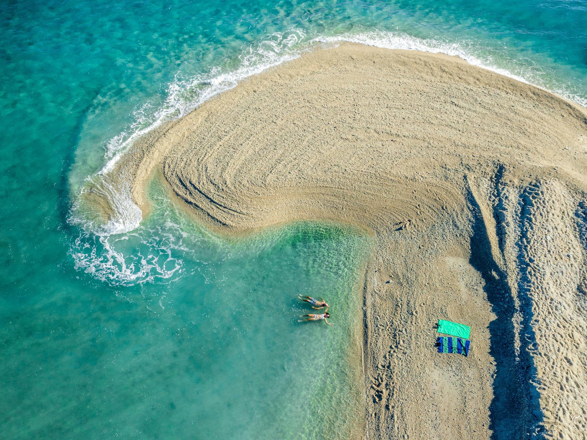 Ariel view of a sand spit curling into tropical blue waters with two swimmers.