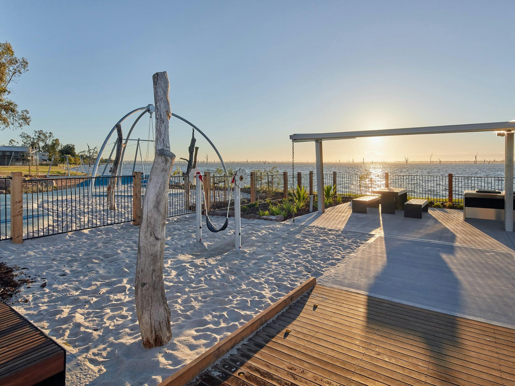 Play equipment in white sand, a wooden boardwalk and BBQ seating overlooking Lake Mulwala.