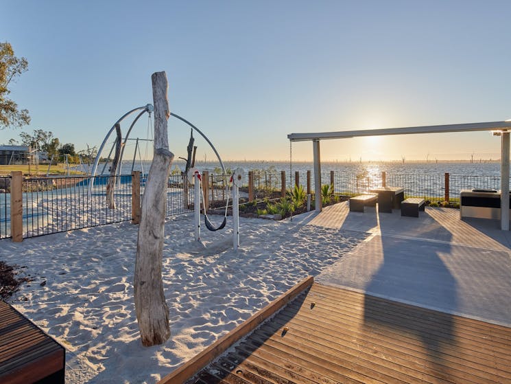 Play equipment in white sand, a wooden boardwalk and BBQ seating overlooking Lake Mulwala.