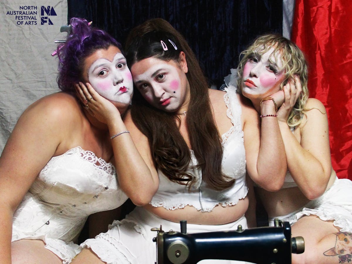 3 young women in white corsets, bloomers and clown makeup lean on each other surrounded by material.