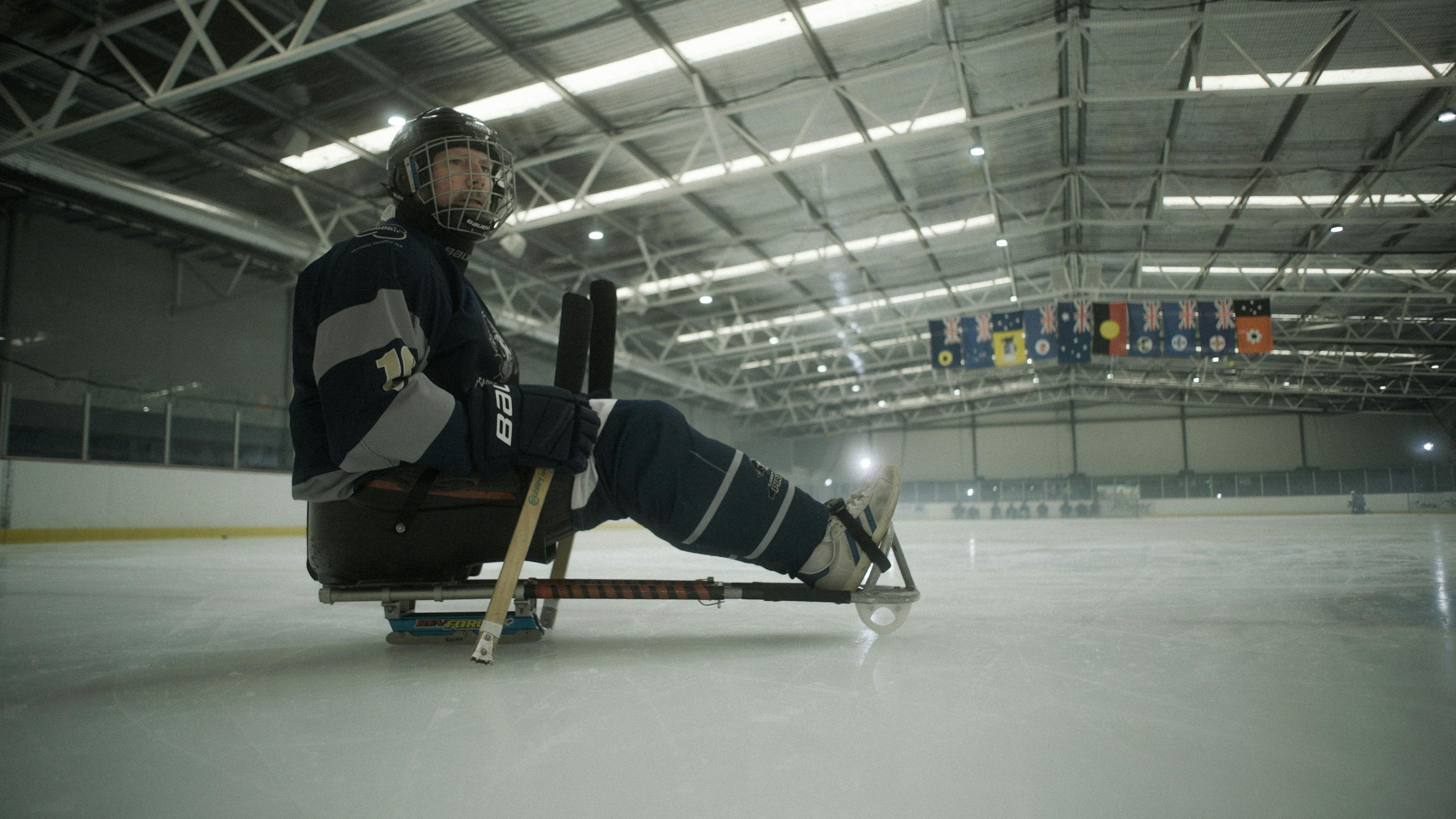 a para ice hockey player sits in their sled, their hockey sticks balancing them