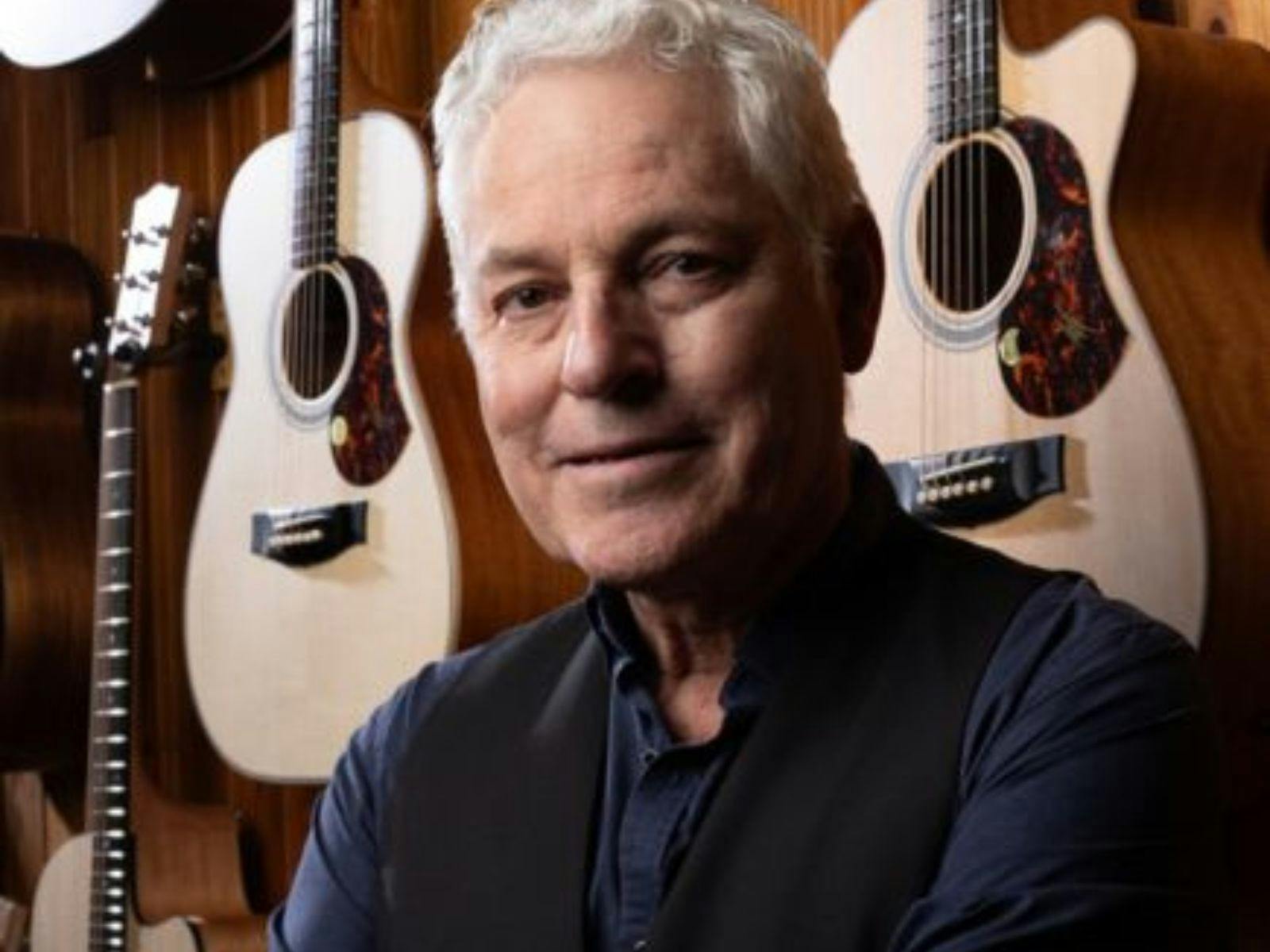 A grey haired man with a wall behind him with guitars on it