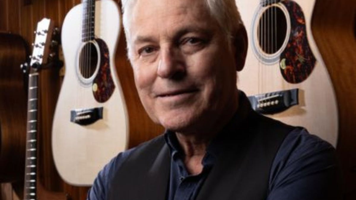 A grey haired man with a wall behind him with guitars on it