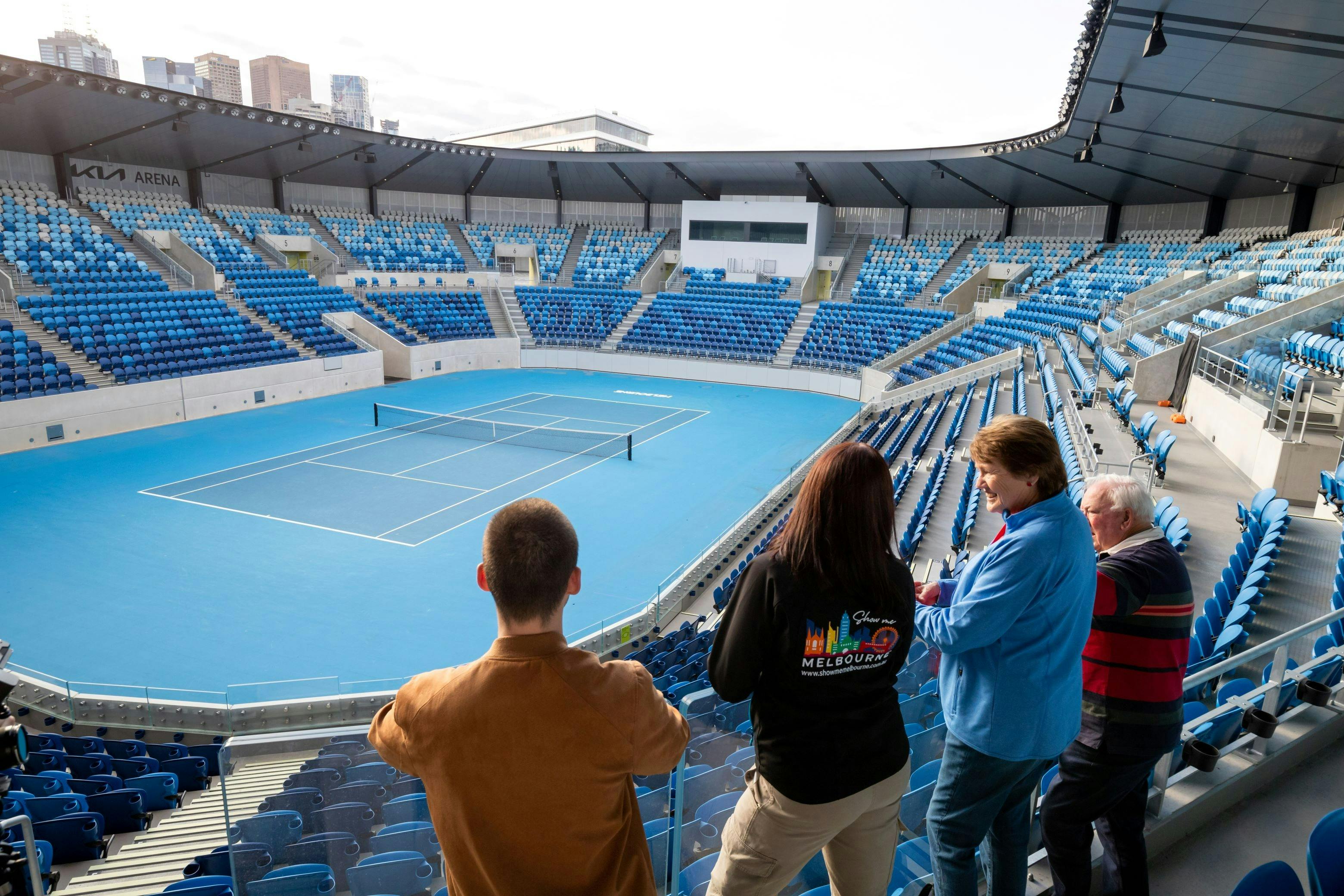 A group of four people are looking at a blue tennis court from the stands.  One is a tour guide.