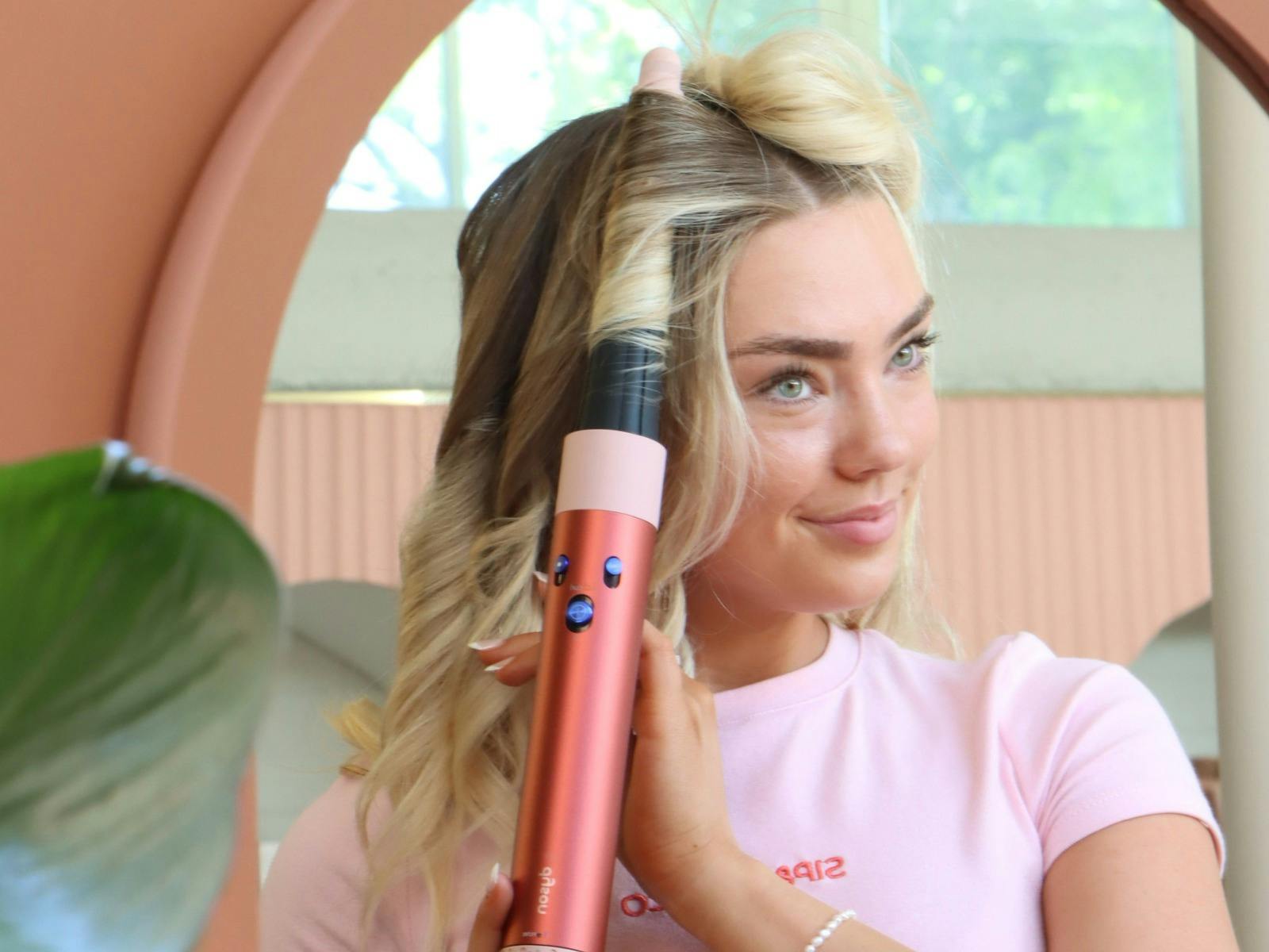 Smiling woman curling her hair with a styling tool while looking in the mirror at a beauty studio.