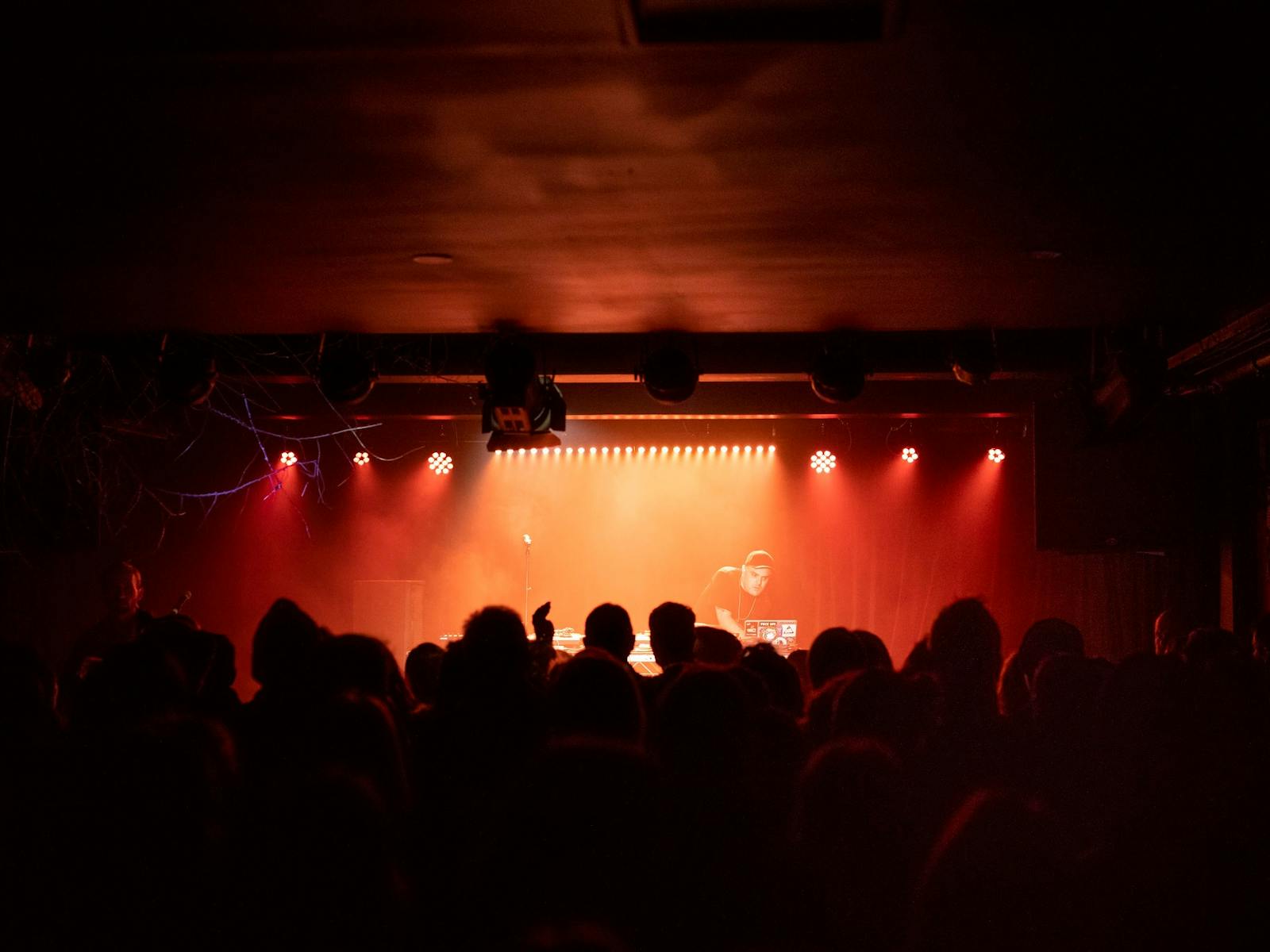 A DJ plays decks on an upraised stage with orange lighting, a crowd looks on