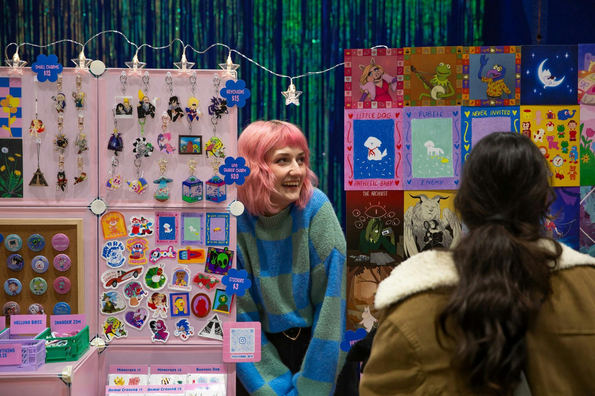 Vendor with pink hair chatting to customer at colourful art stall