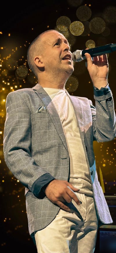 A man in a jacket stands with a microphone with glitter and drums in the background
