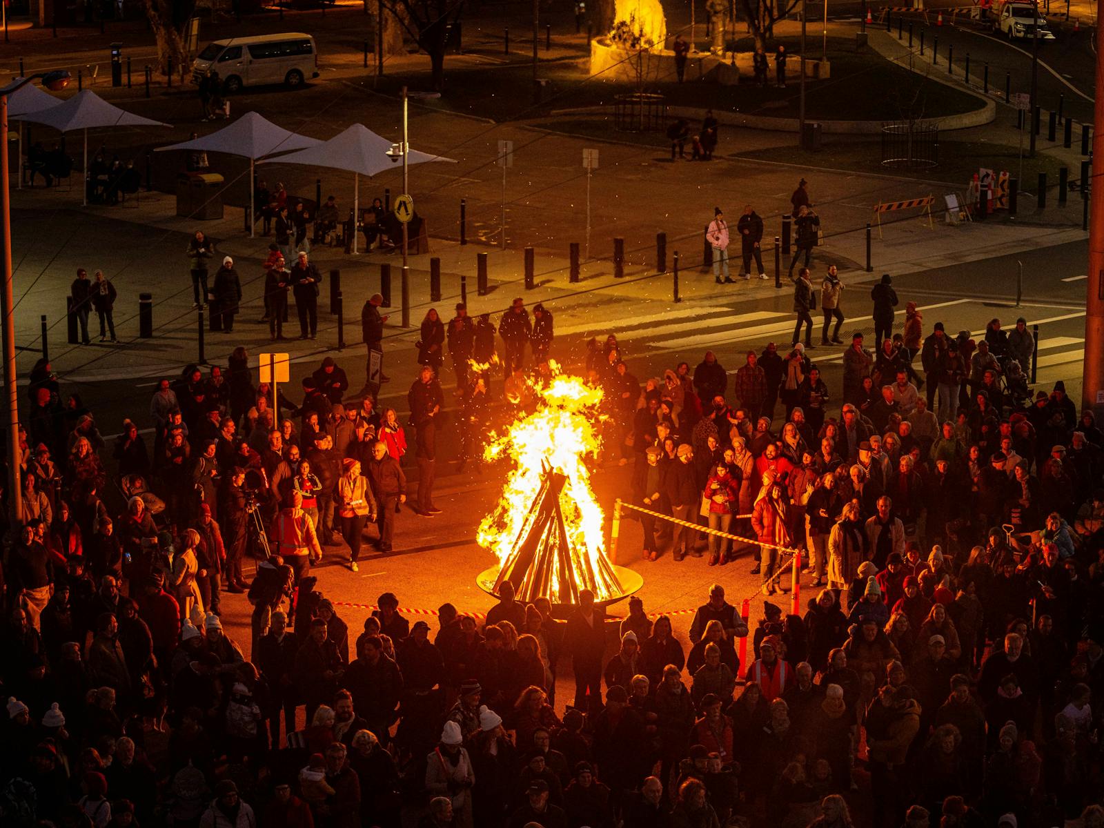 A colour photograph at nighttime with a large bonfire shining bright with people circled around