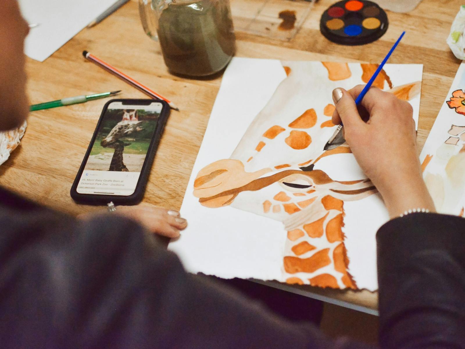 A person painting a picture of an adult and juvenile giraffe with watercolour