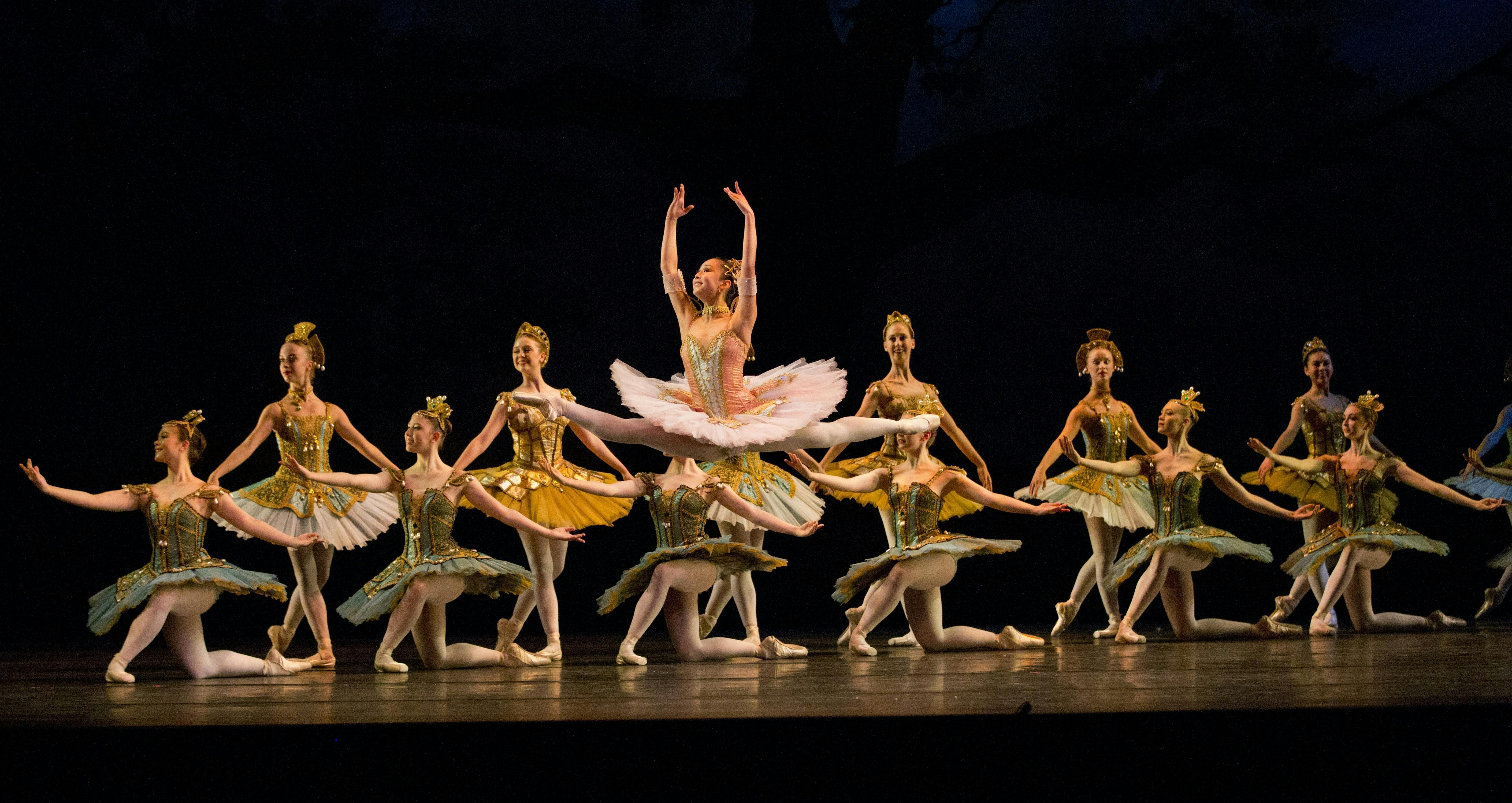 Ballerinas in a line on a stage. One dancer in center is mid jump. They all wear gold/white tutus.