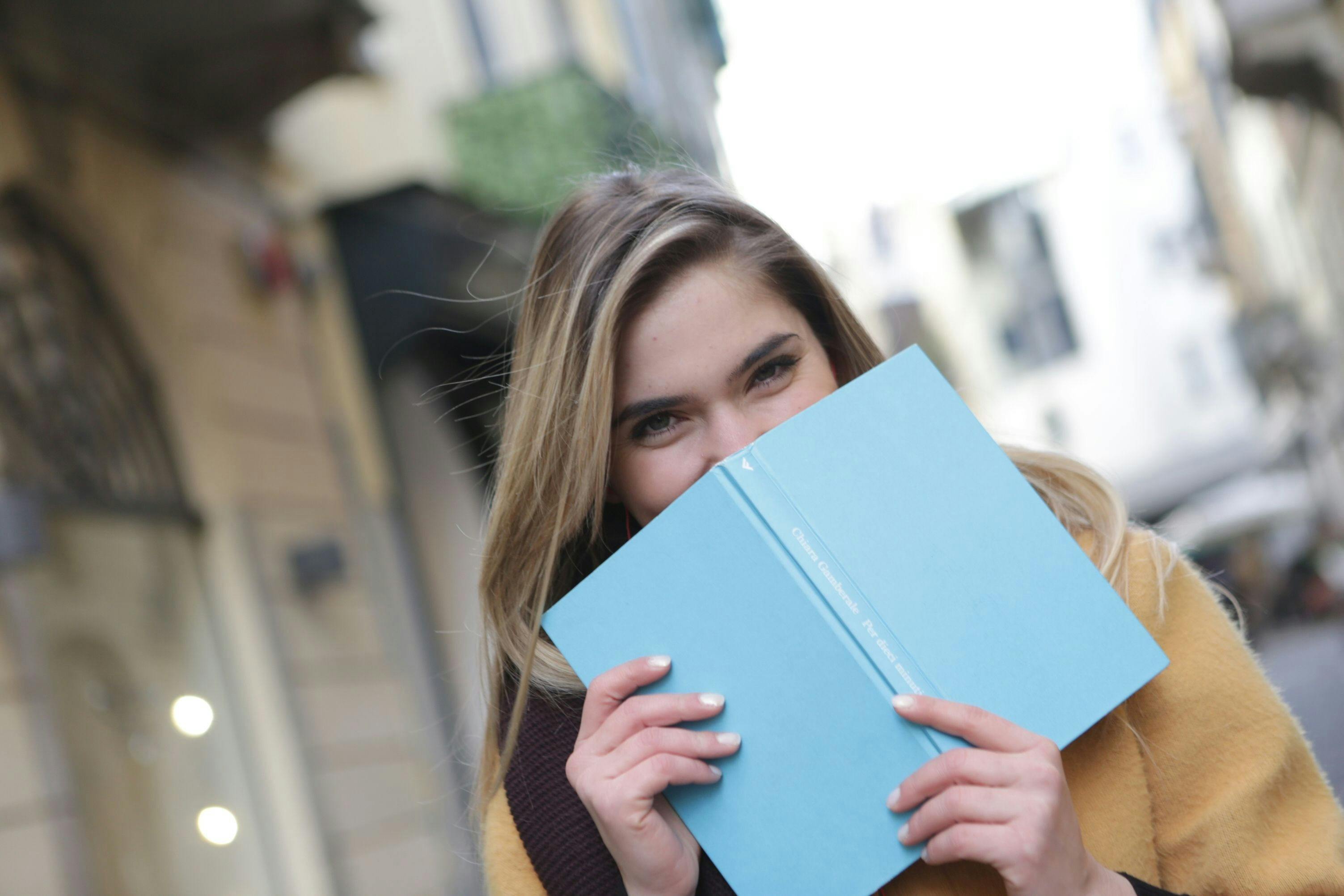 Woman with blonde hari holding up a blue book to half cover her face