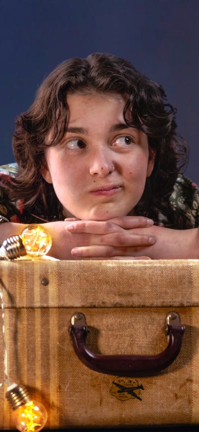 A young woman with short dark curls rests her chin on her hands on top of a brown vintage suitcase.